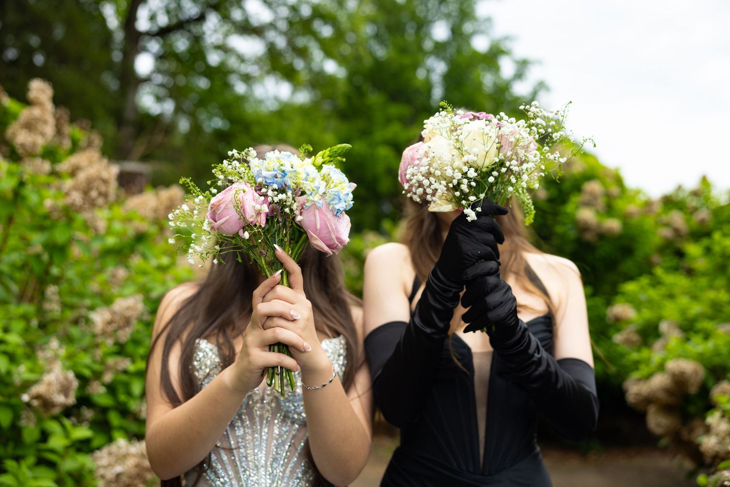 Two young women in formal dresses hold floral bouquets covering their faces outdoors near blooming hydrangeas.