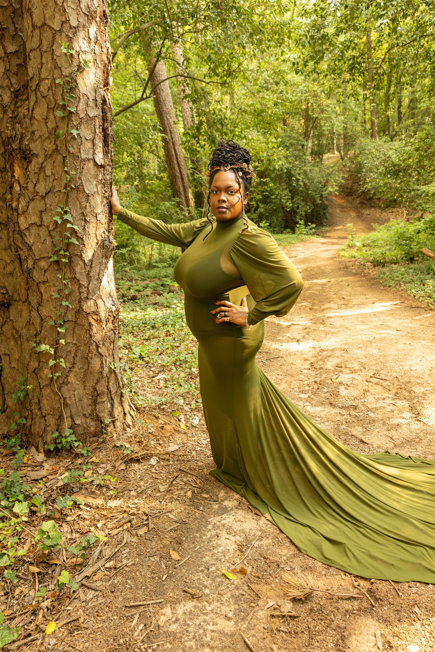 Pregnant woman in elegant olive green gown poses by a tree on a forest path.