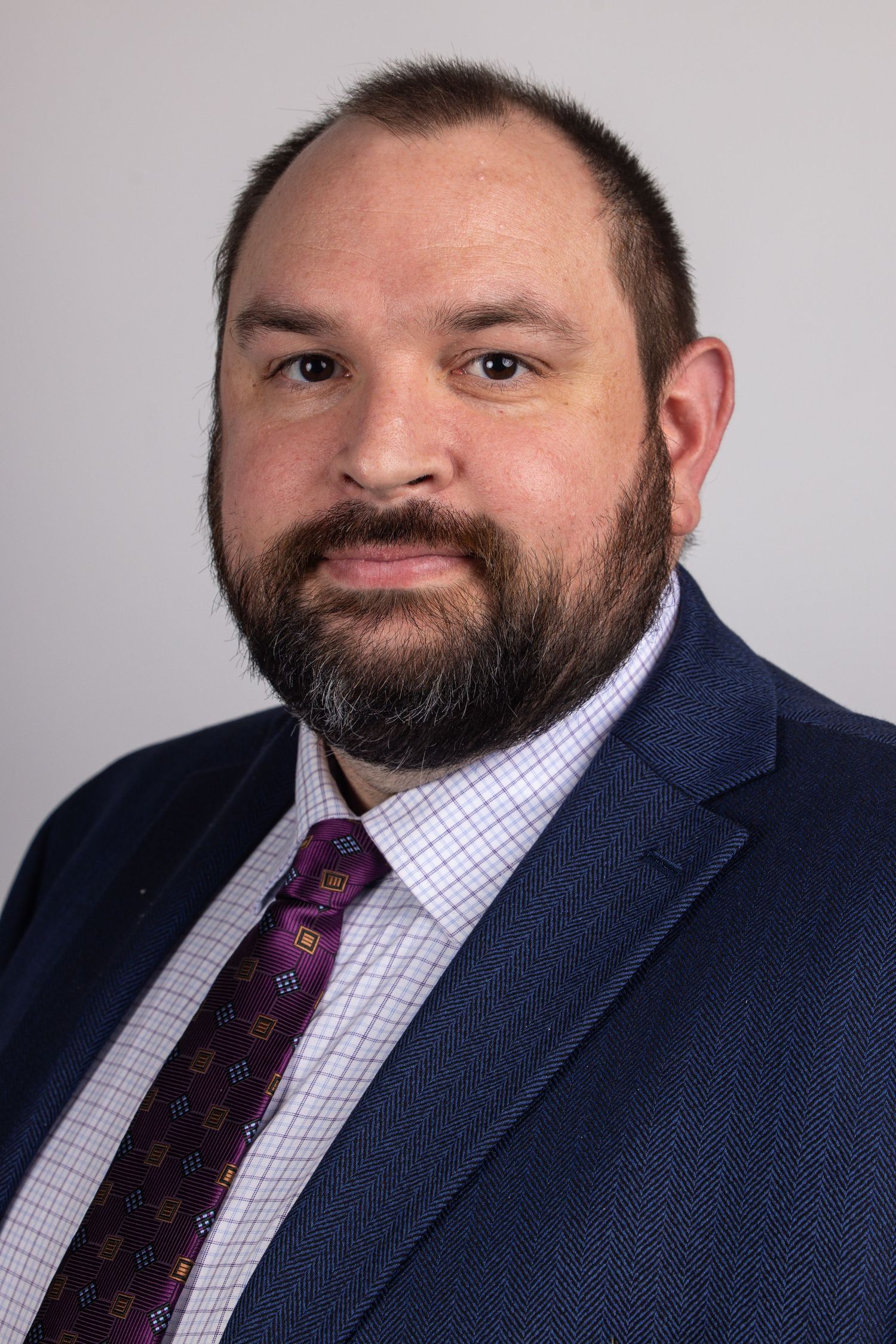 Professional headshot of a bearded man in a navy blue suit with a burgundy patterned tie.