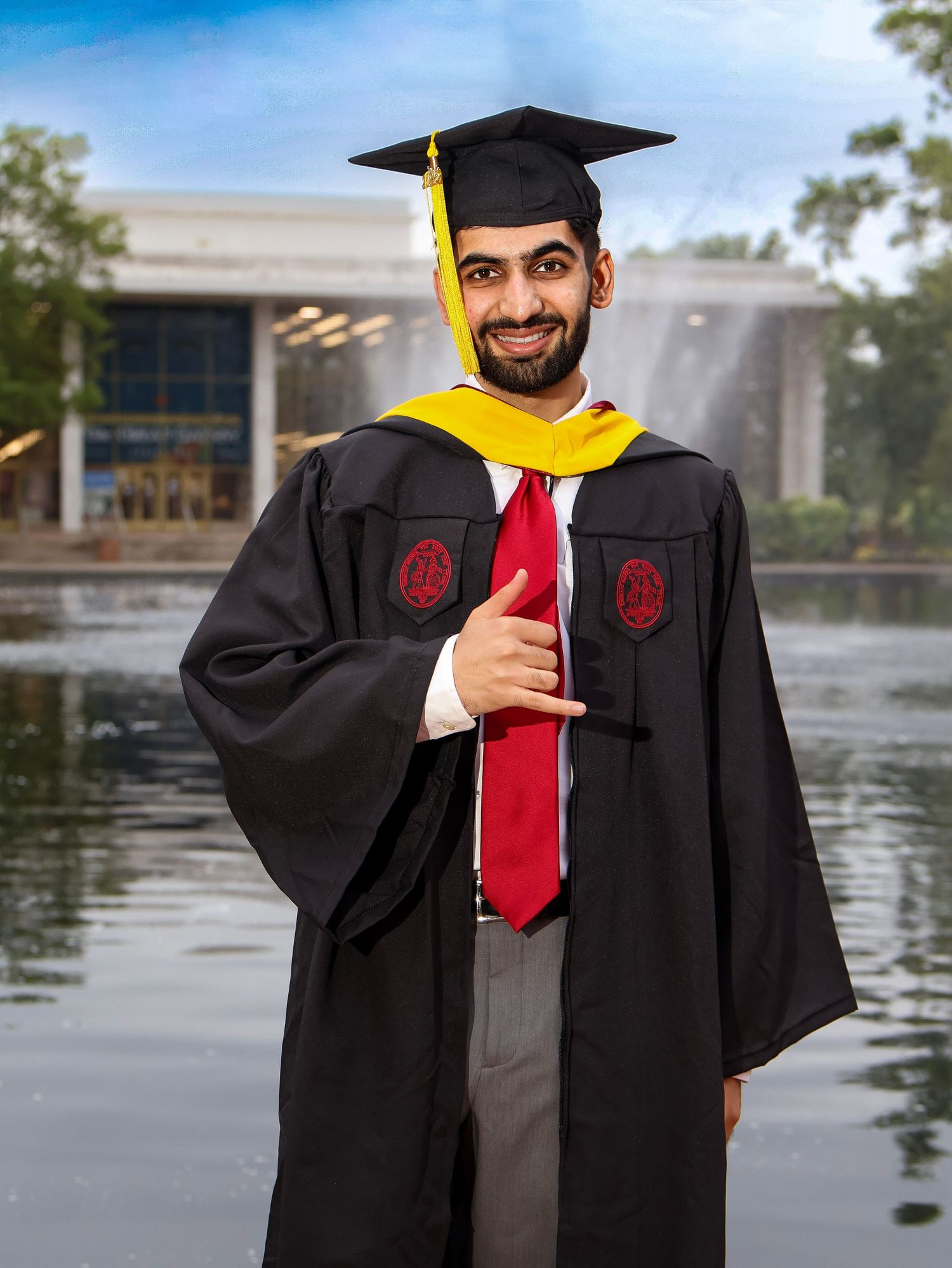 Graduate in black cap and gown with yellow stole poses by fountain.