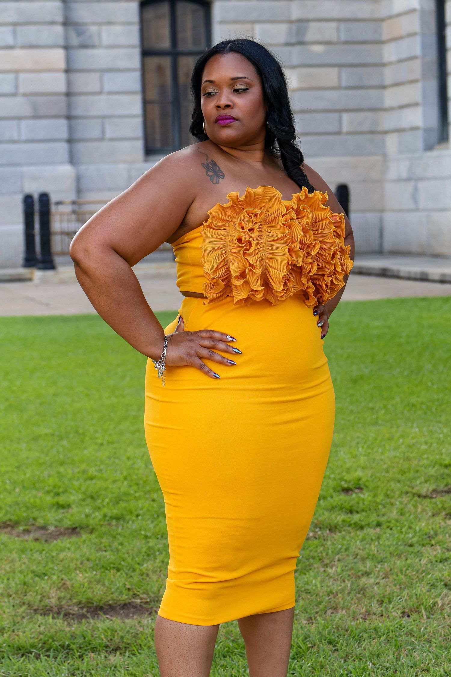 Model in bright yellow ruffle-detailed bodycon dress poses against stone building backdrop.
