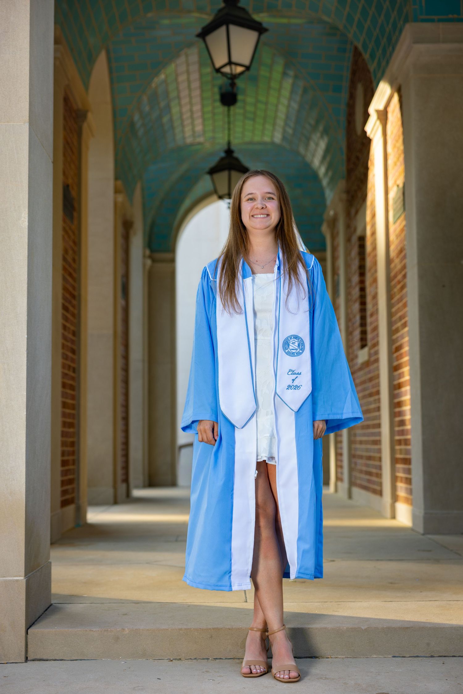 Young woman smiling in light blue graduation gown and stole, standing in a brick archway colonnade.