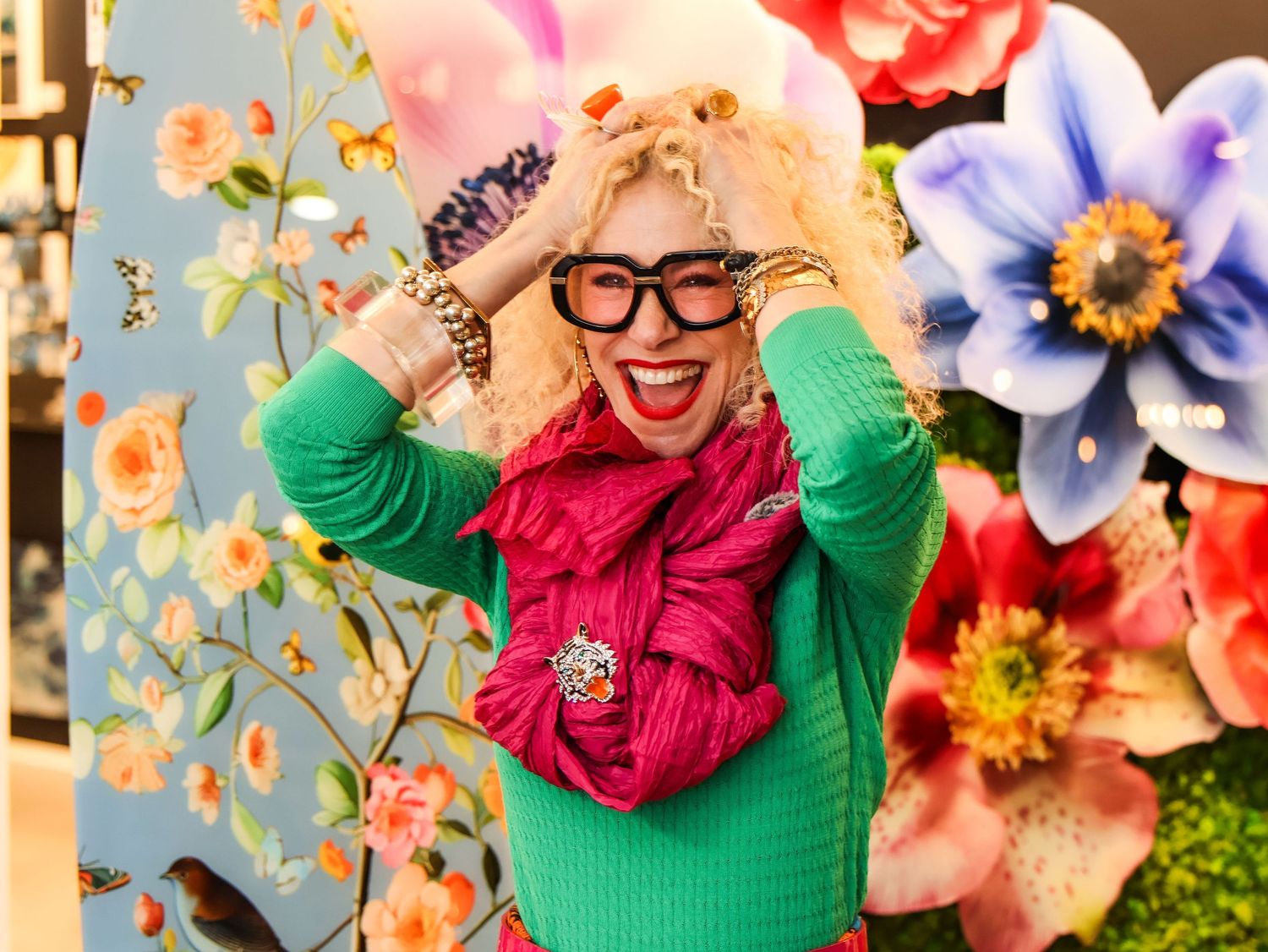Person in green sweater and pink scarf posing against floral backdrop.