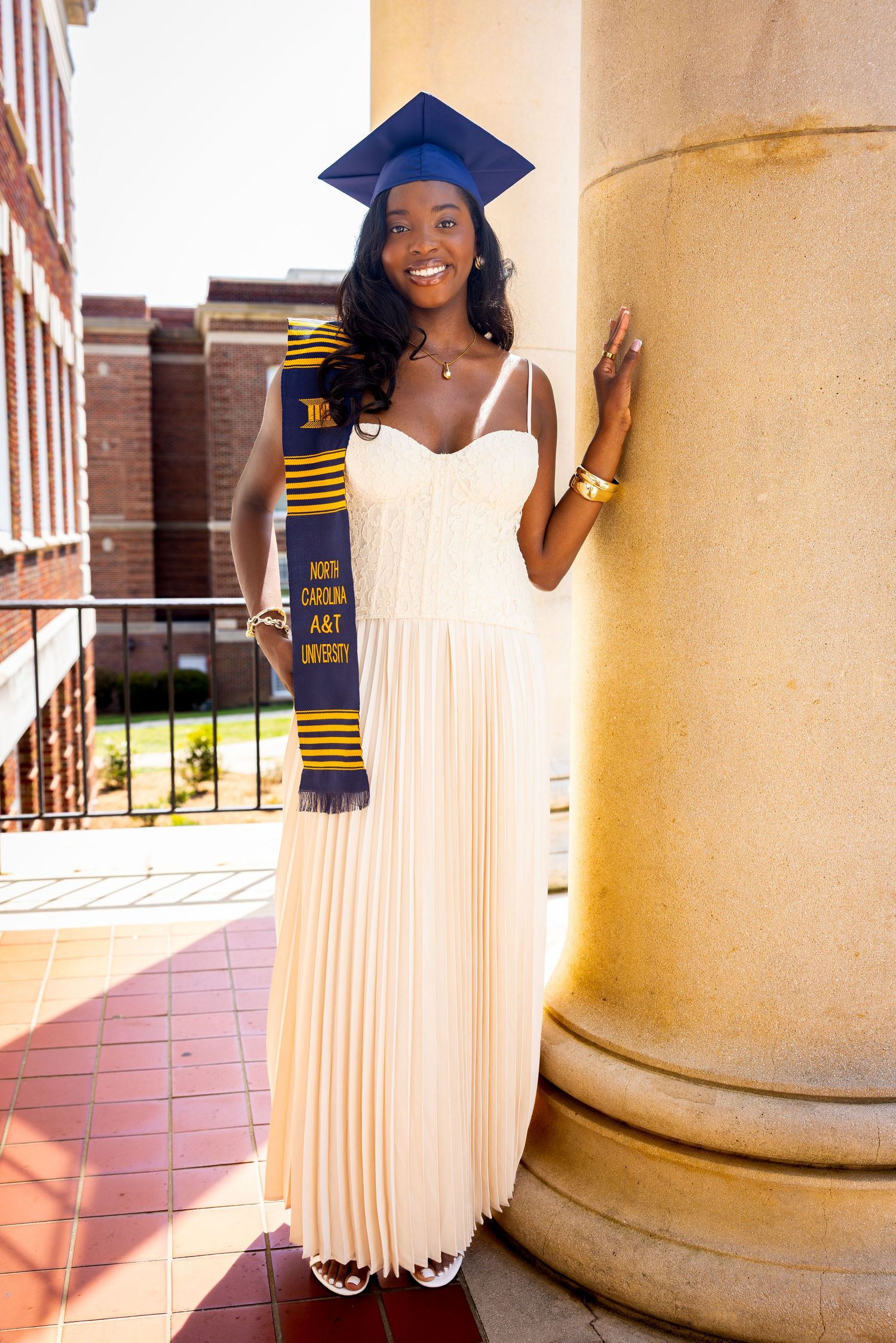 Smiling graduate in white gown and blue cap holds North Carolina A&T University stole beside a column.