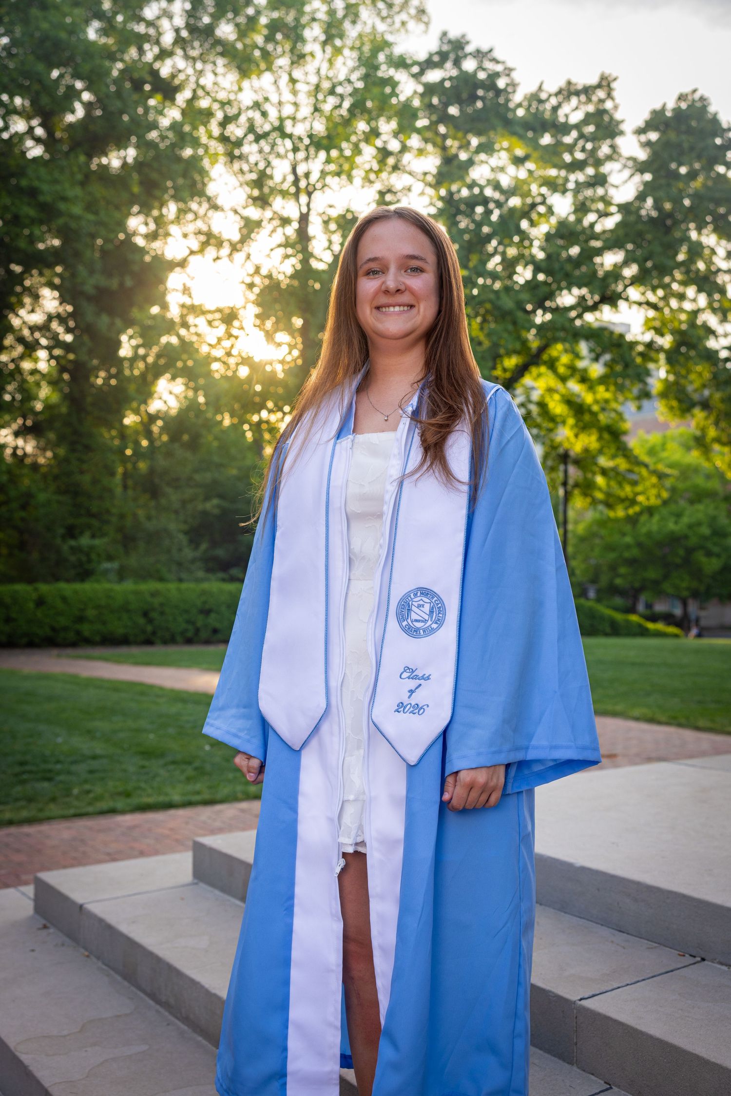 Young woman in light blue graduation gown and white stole smiling outdoors at sunset on campus steps.