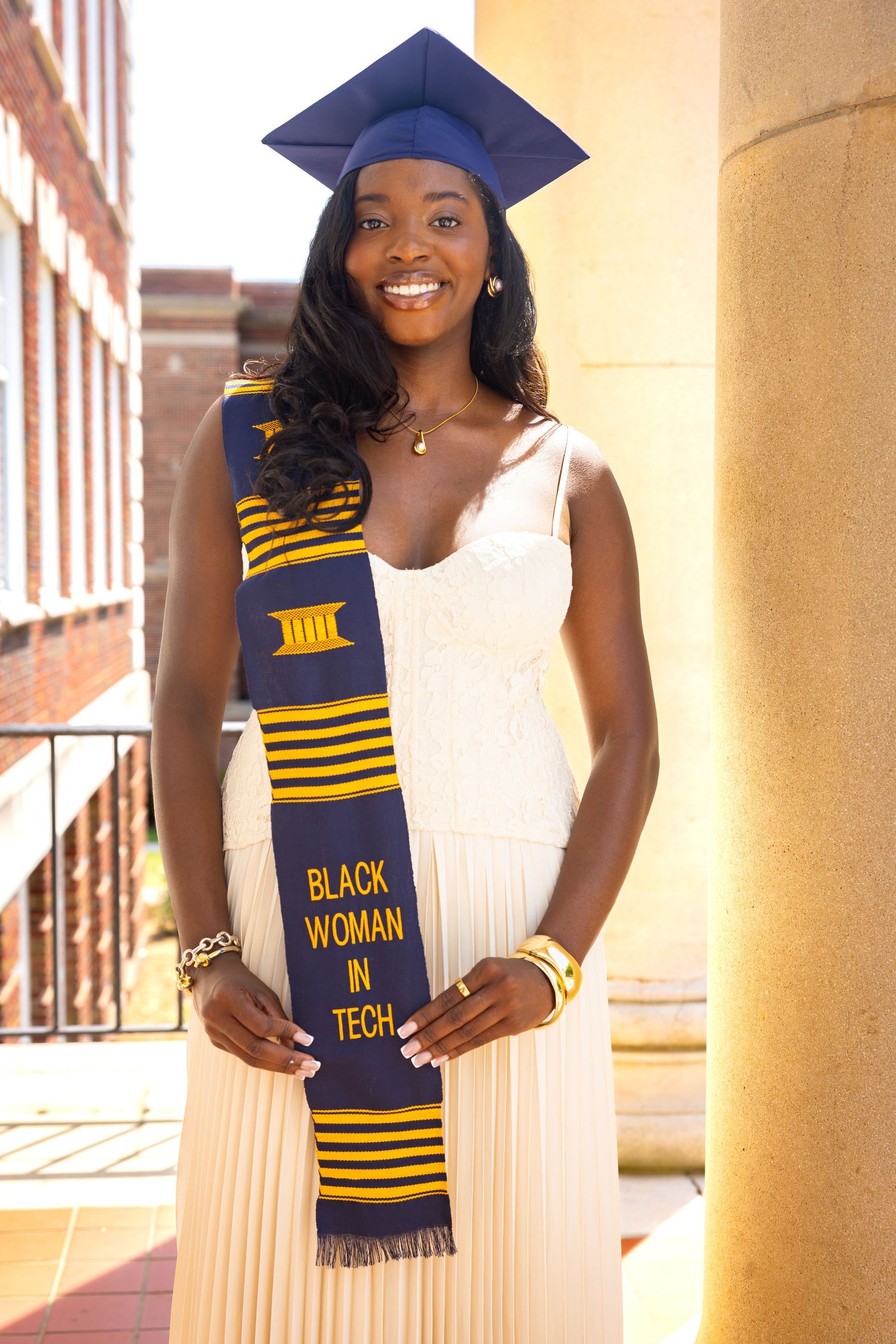 Smiling graduate in cap and white dress wearing a Black Woman in Tech kente stole, posing by a column.