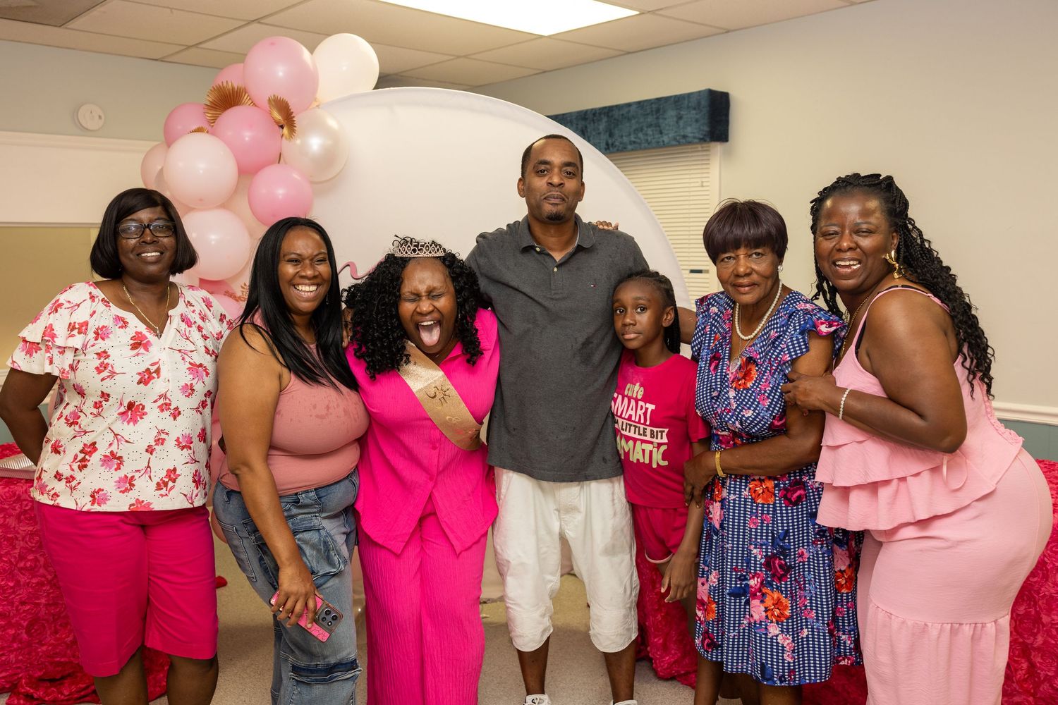 Group of people in pink outfits celebrating at a party with balloon decorations in the background.