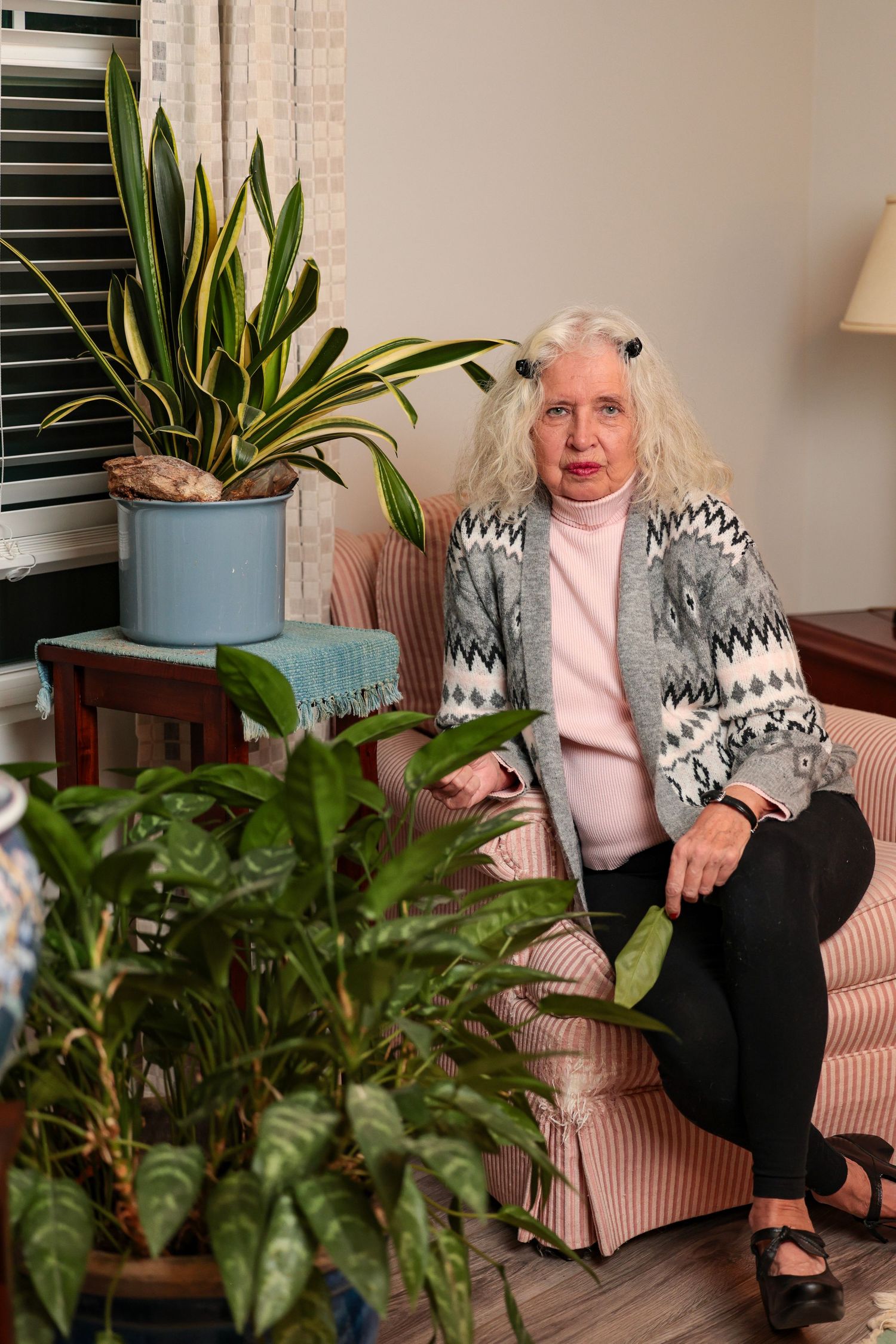 Person sitting on pink couch surrounded by houseplants wearing gray patterned sweater and pink top.