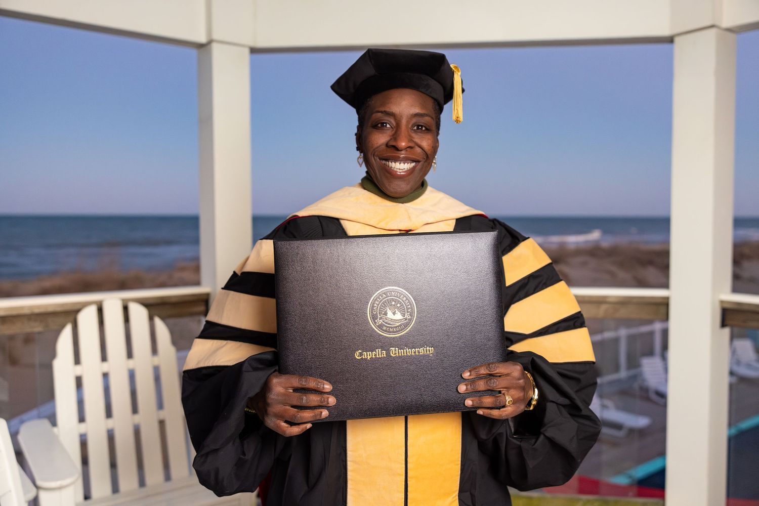 Woman in doctoral graduation gown and cap holds Capella University diploma by the ocean.