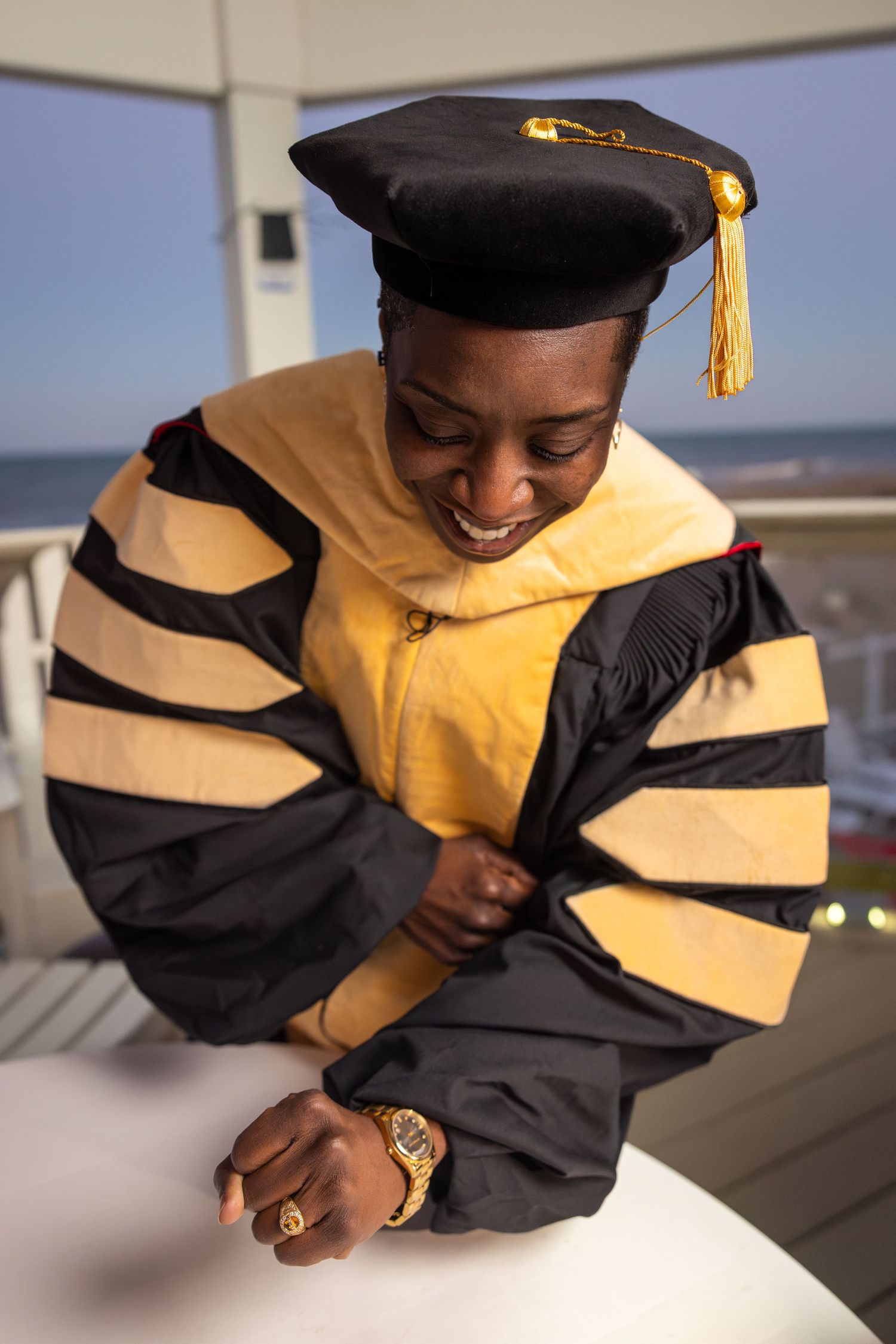Smiling graduate in black and gold doctoral regalia and cap with tassel, looking down outdoors near ocean.