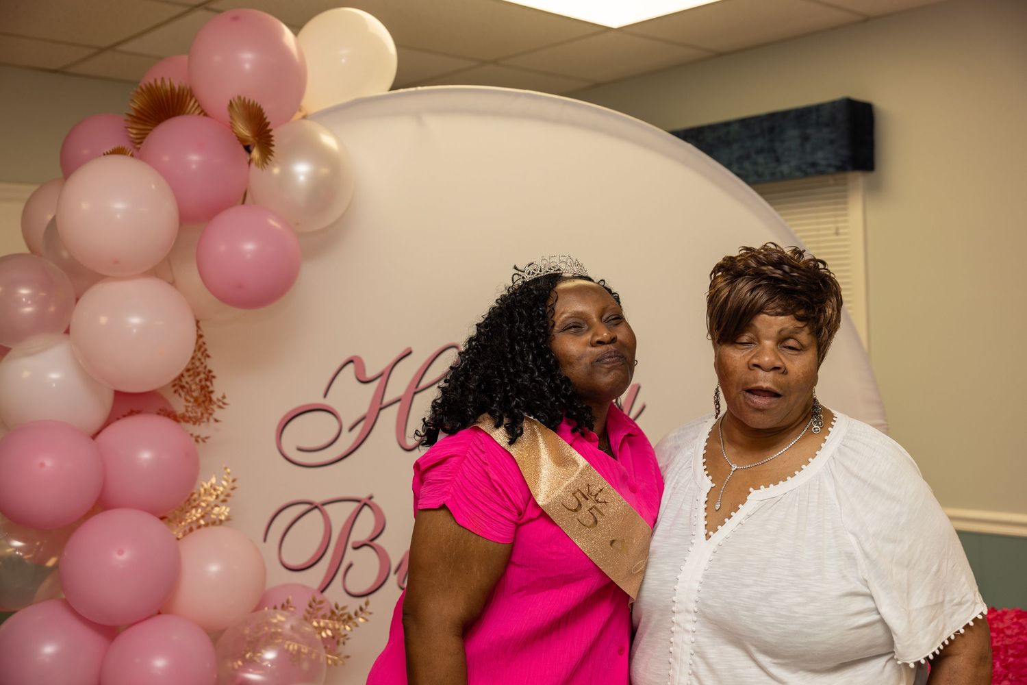 Two women posing together at a pink and white balloon decorated party backdrop celebration event.