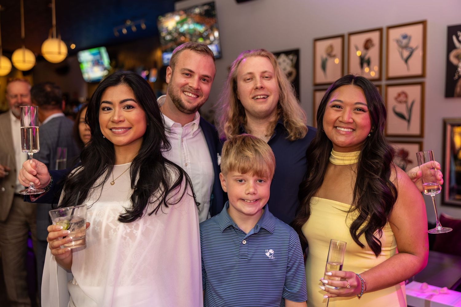 Group of five people smiling at a social gathering, holding champagne glasses in a warmly lit restaurant.