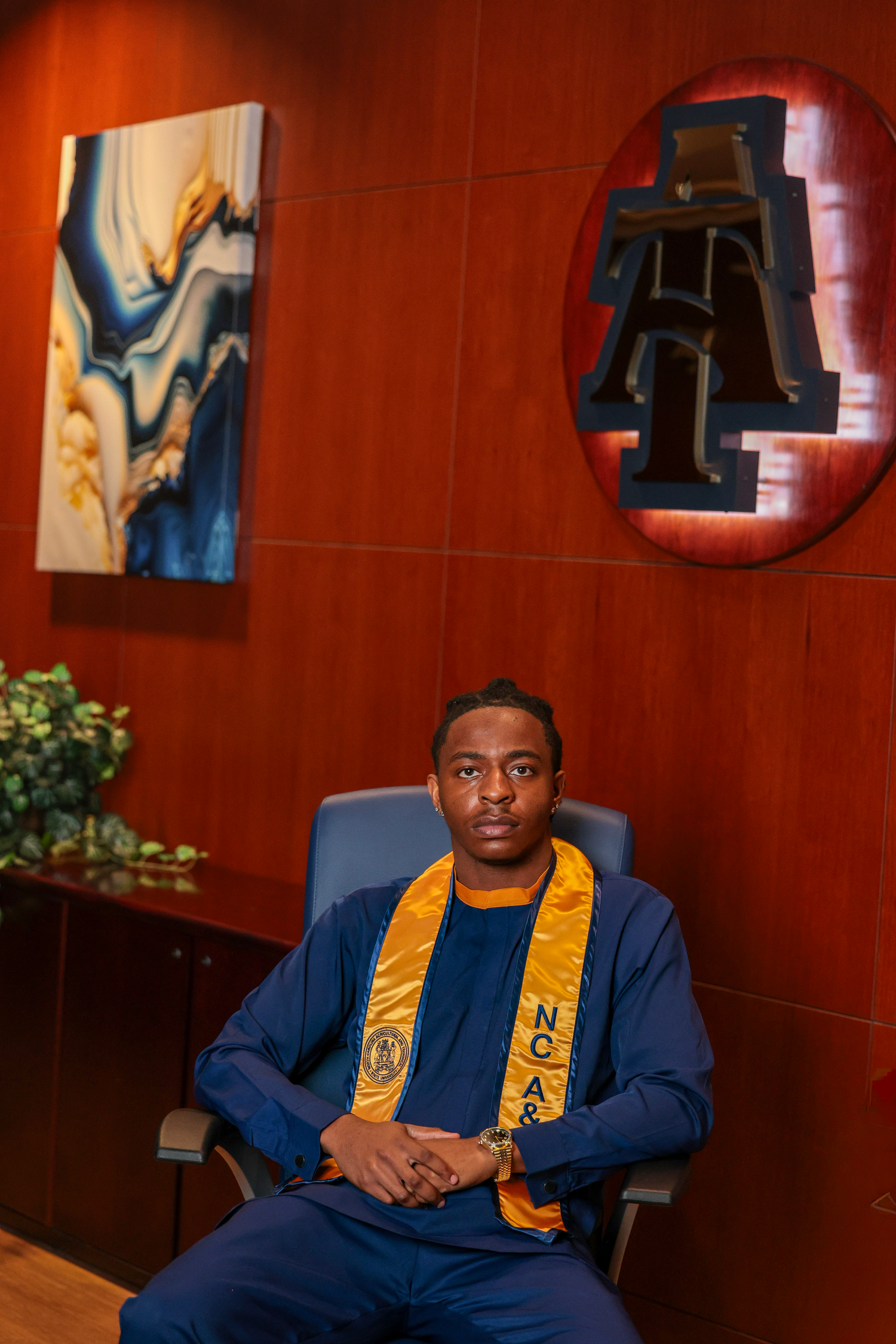 Graduate in academic regalia sits for portrait against red paneled wall.