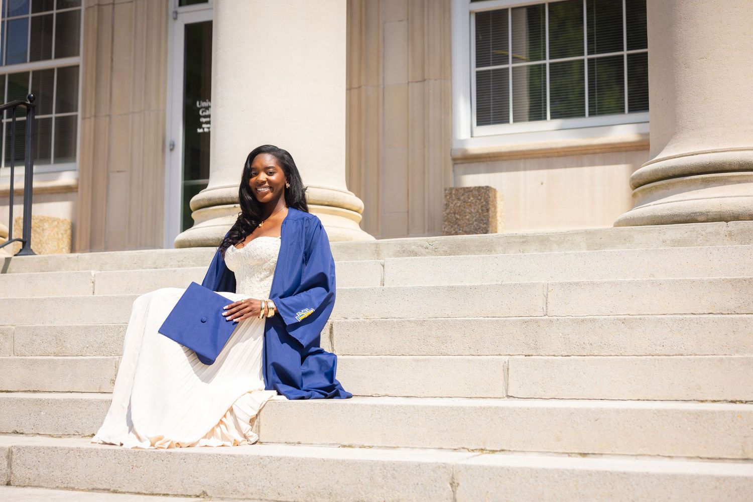 A smiling graduate in a blue cap and gown sits on stone steps outside a classical building, holding her diploma.