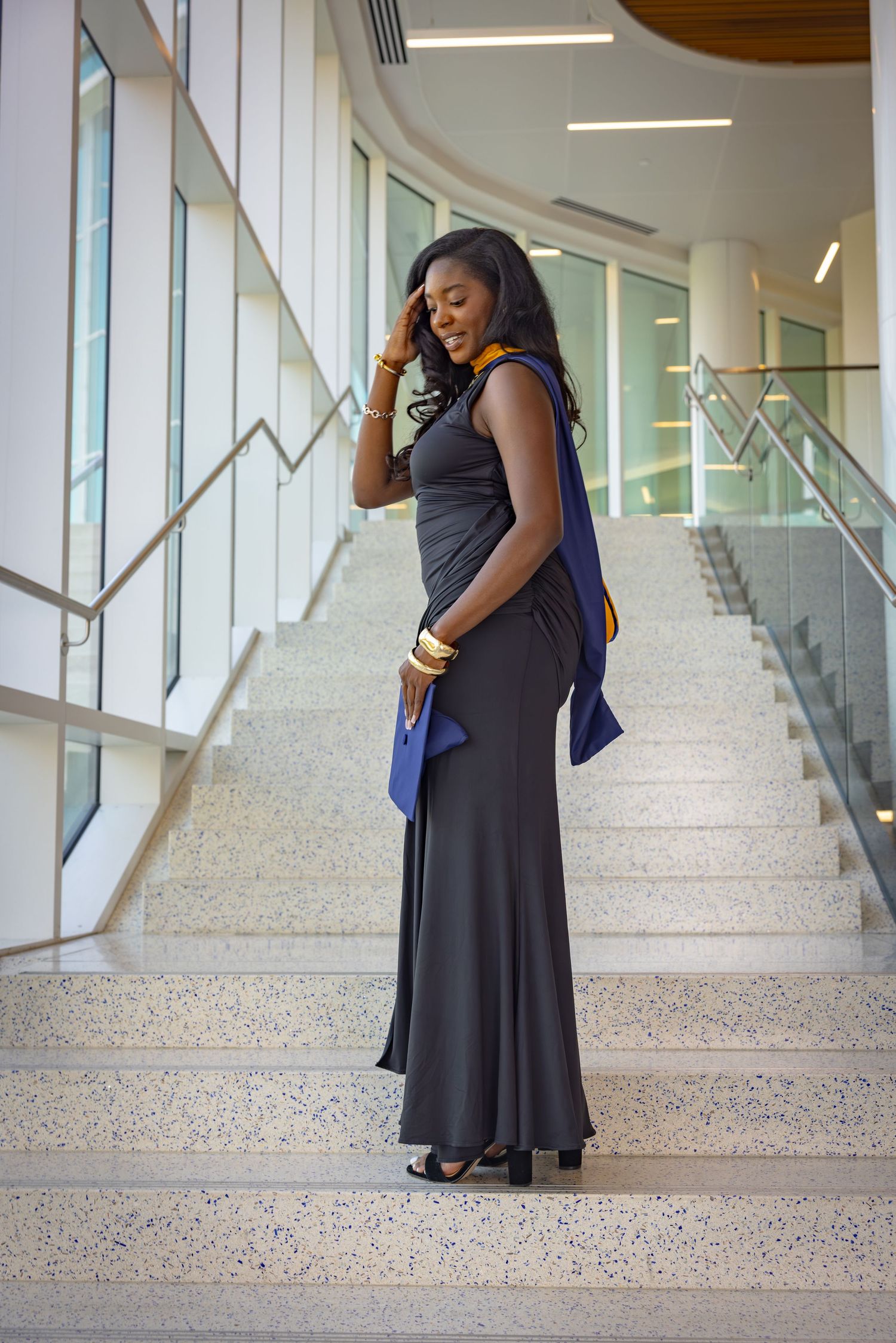 Elegant woman in black maxi dress holding blue clutch on modern building stairs with gold accessories.