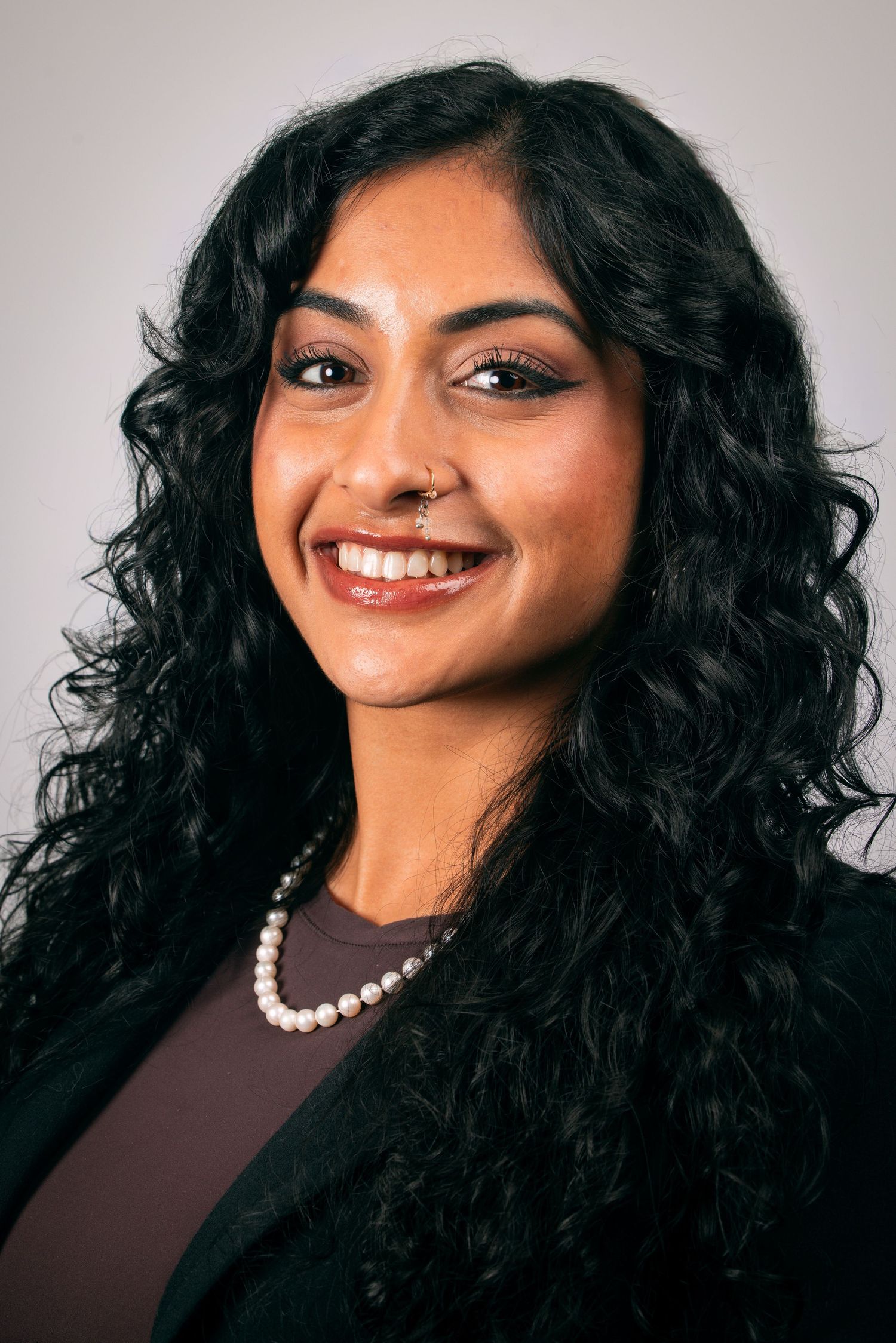 Smiling South Asian woman with curly black hair, nose ring, pearl necklace, and black blazer poses for professional headshot.