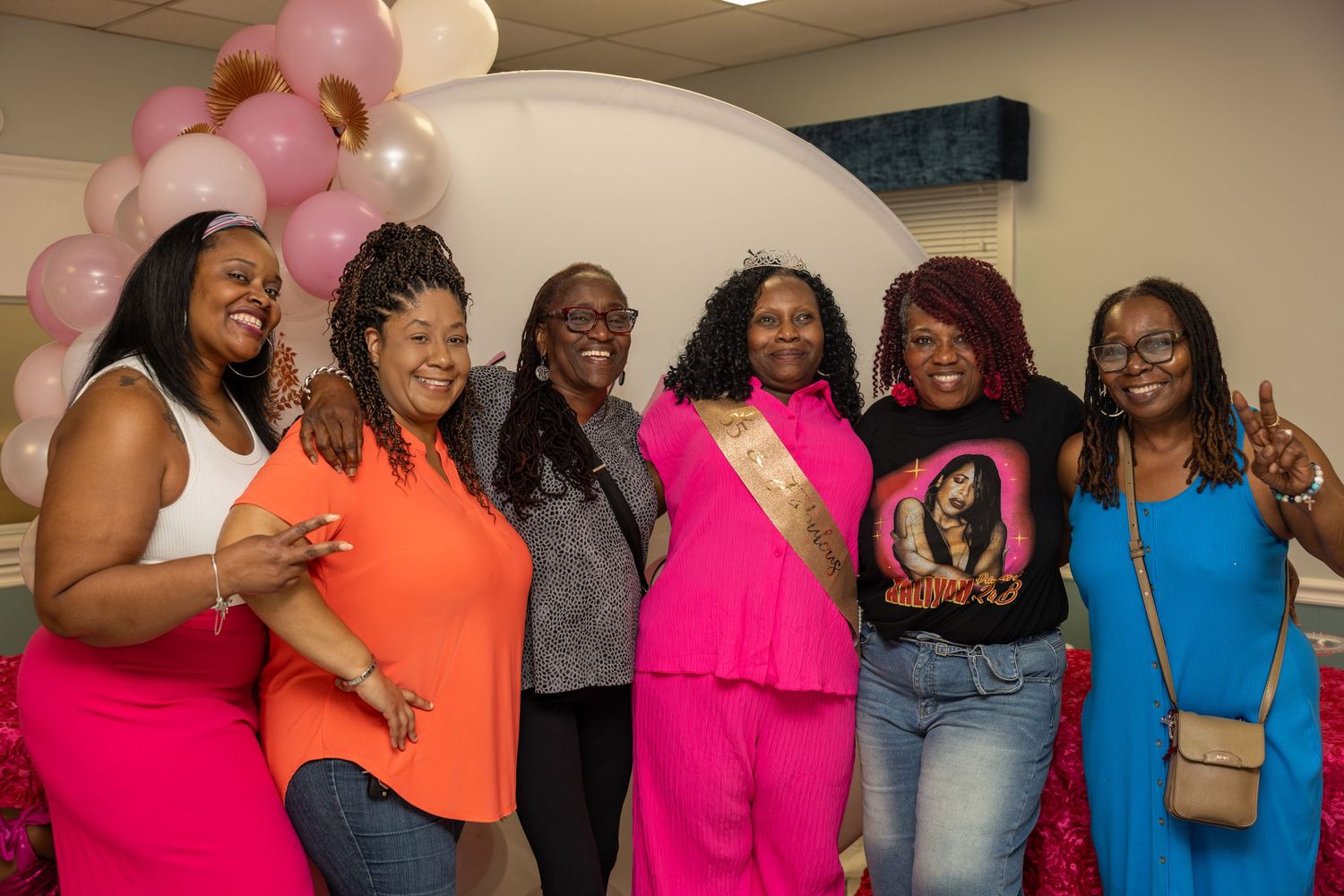 Six women in colorful outfits posing together at a festive party with pink and white balloon arch backdrop.