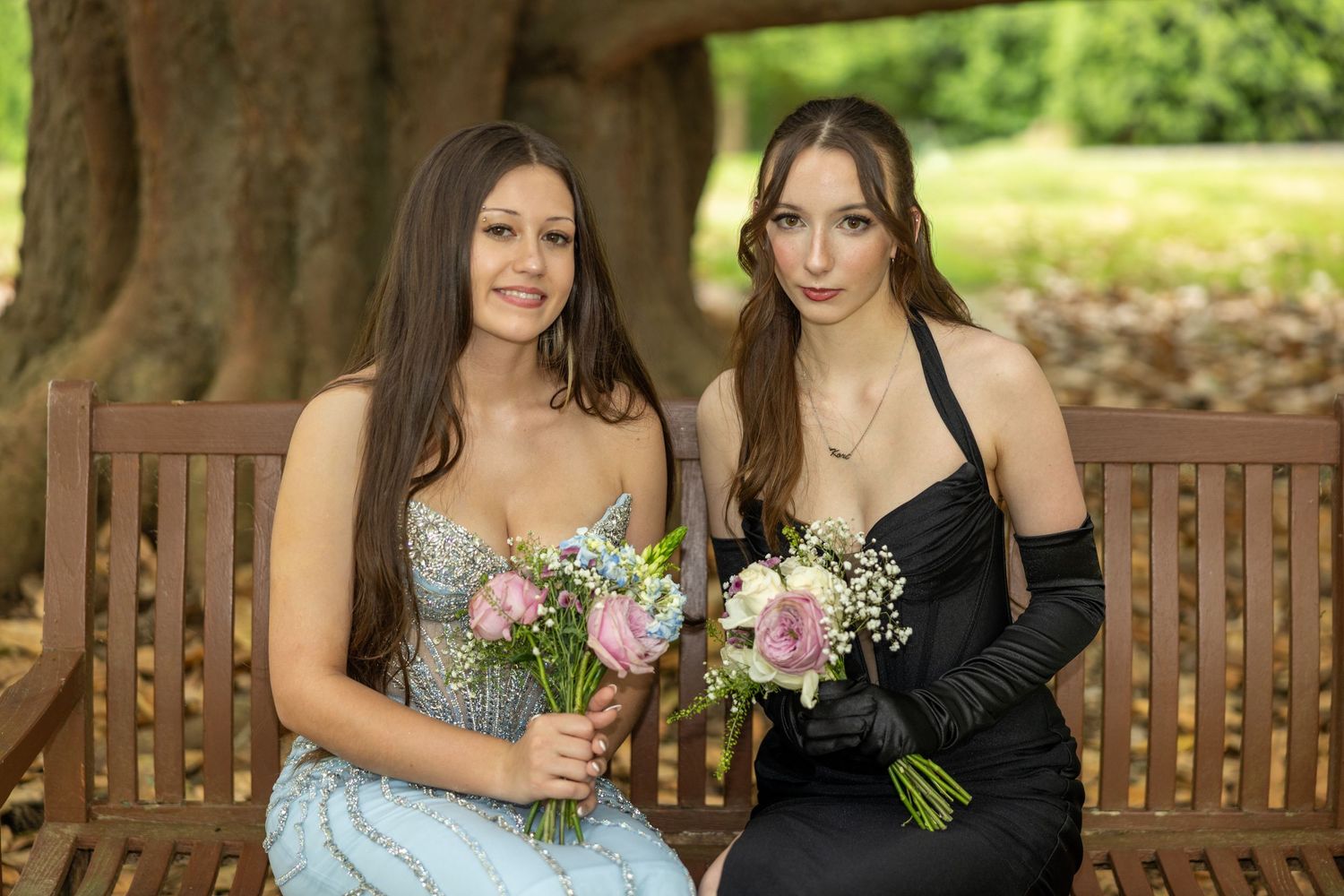 Two elegantly dressed young women holding bouquets sit on a wooden bench outdoors for a formal occasion.