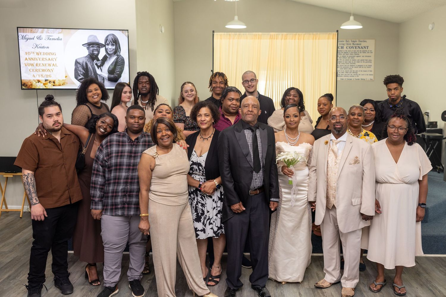 Large family group photo at a wedding celebration, posing together in a bright room with the bride and groom centered.