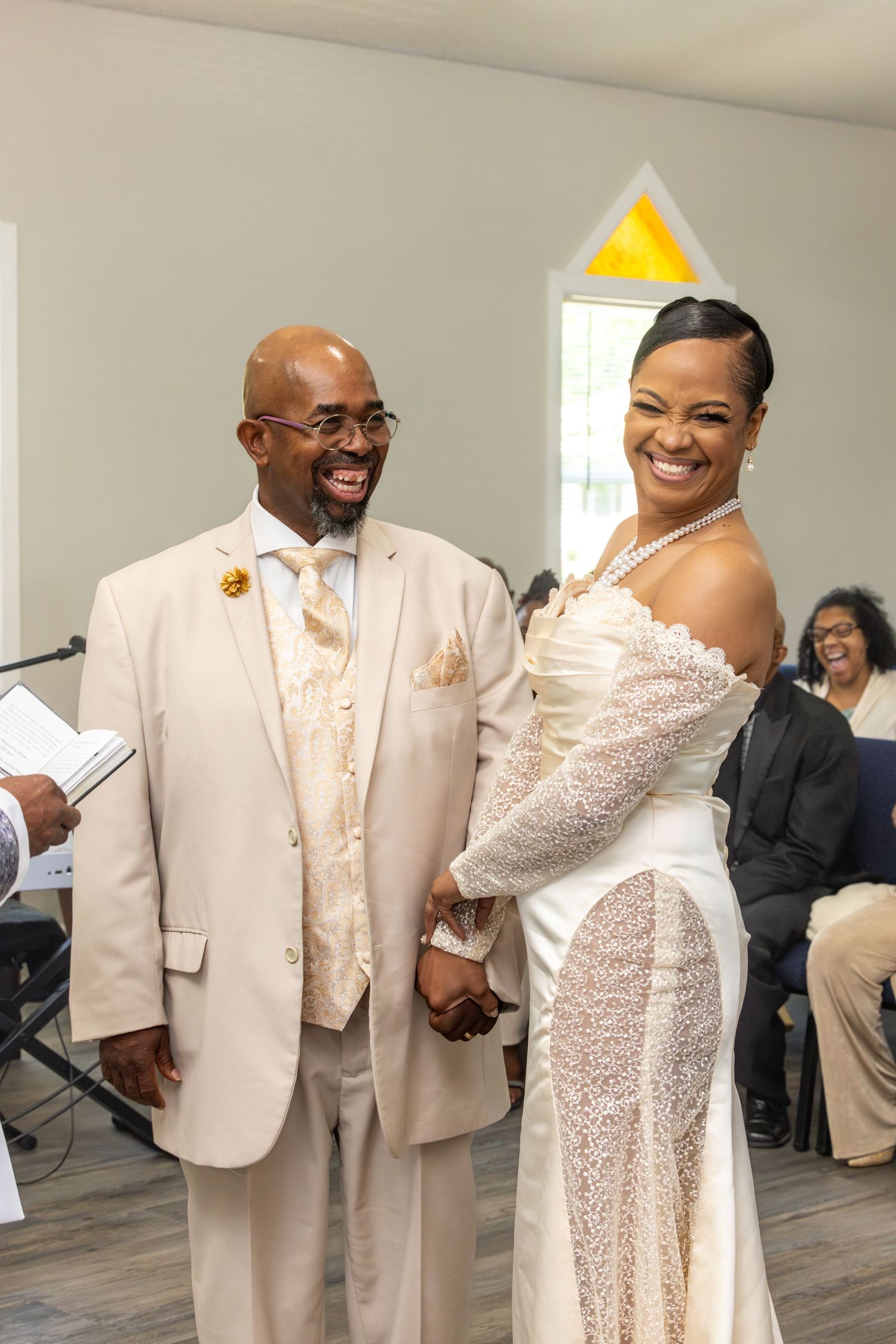 Happy couple holding hands at wedding ceremony, groom in tan suit, bride in elegant lace gown, smiling joyfully.