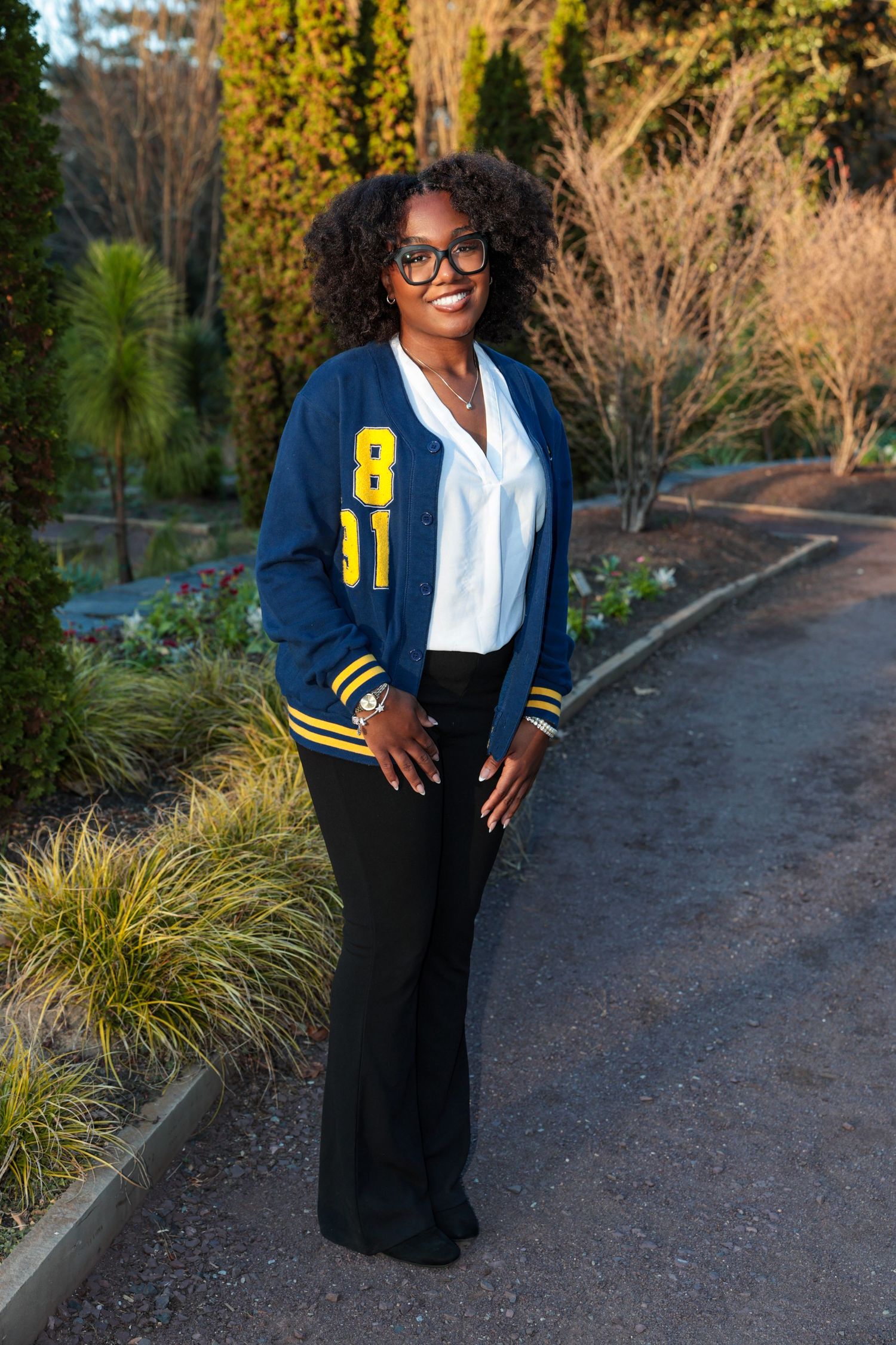 Person in varsity jacket poses outdoors near ornamental grasses.