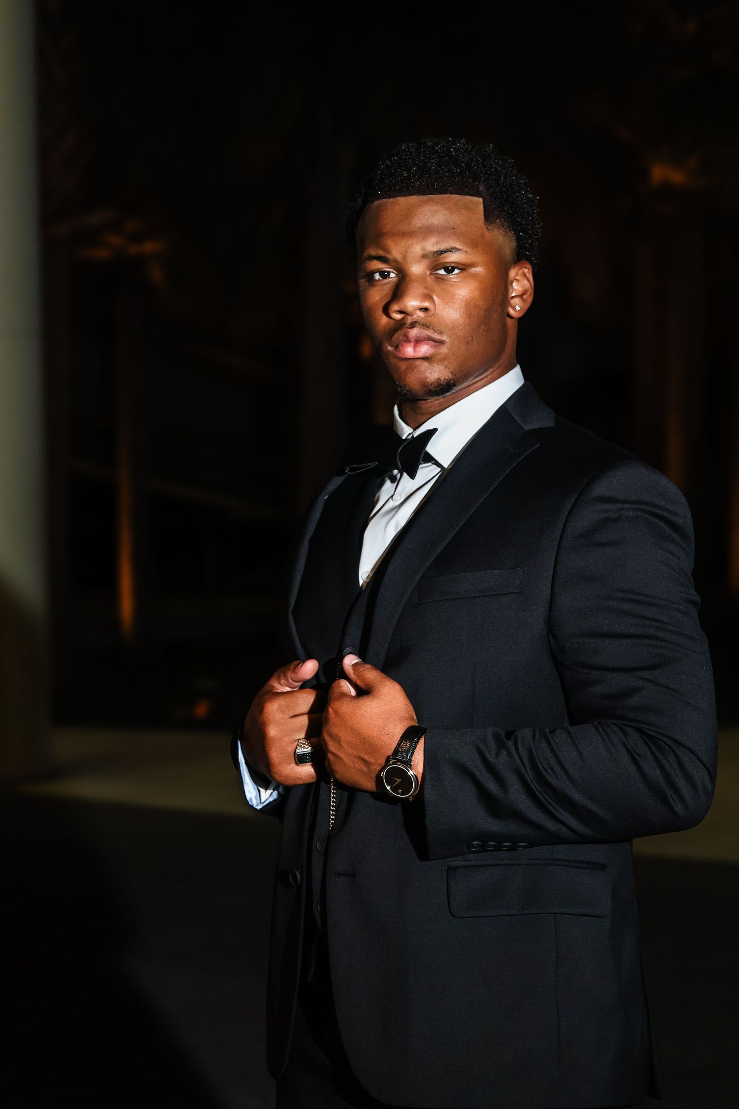 A person in a formal black suit poses dramatically in low-light conditions.
