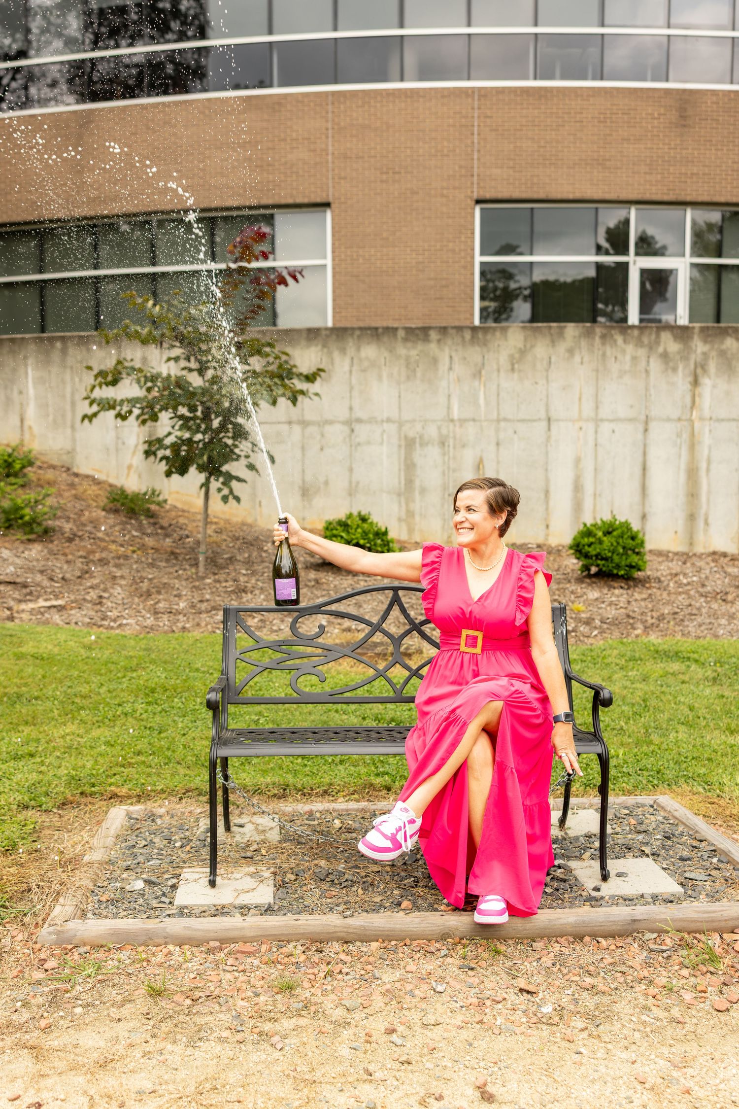 Woman in pink dress spraying champagne while sitting on a black iron bench outside a modern building.