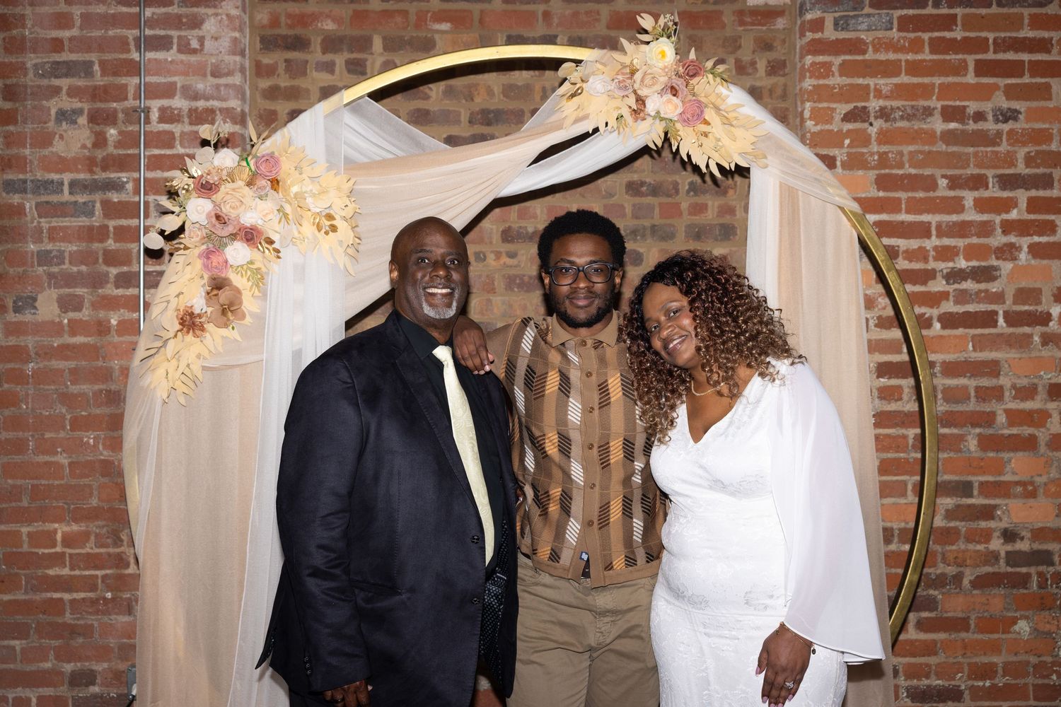 Three people smiling together under a gold circular floral arch with pink flowers at an indoor brick wall venue.