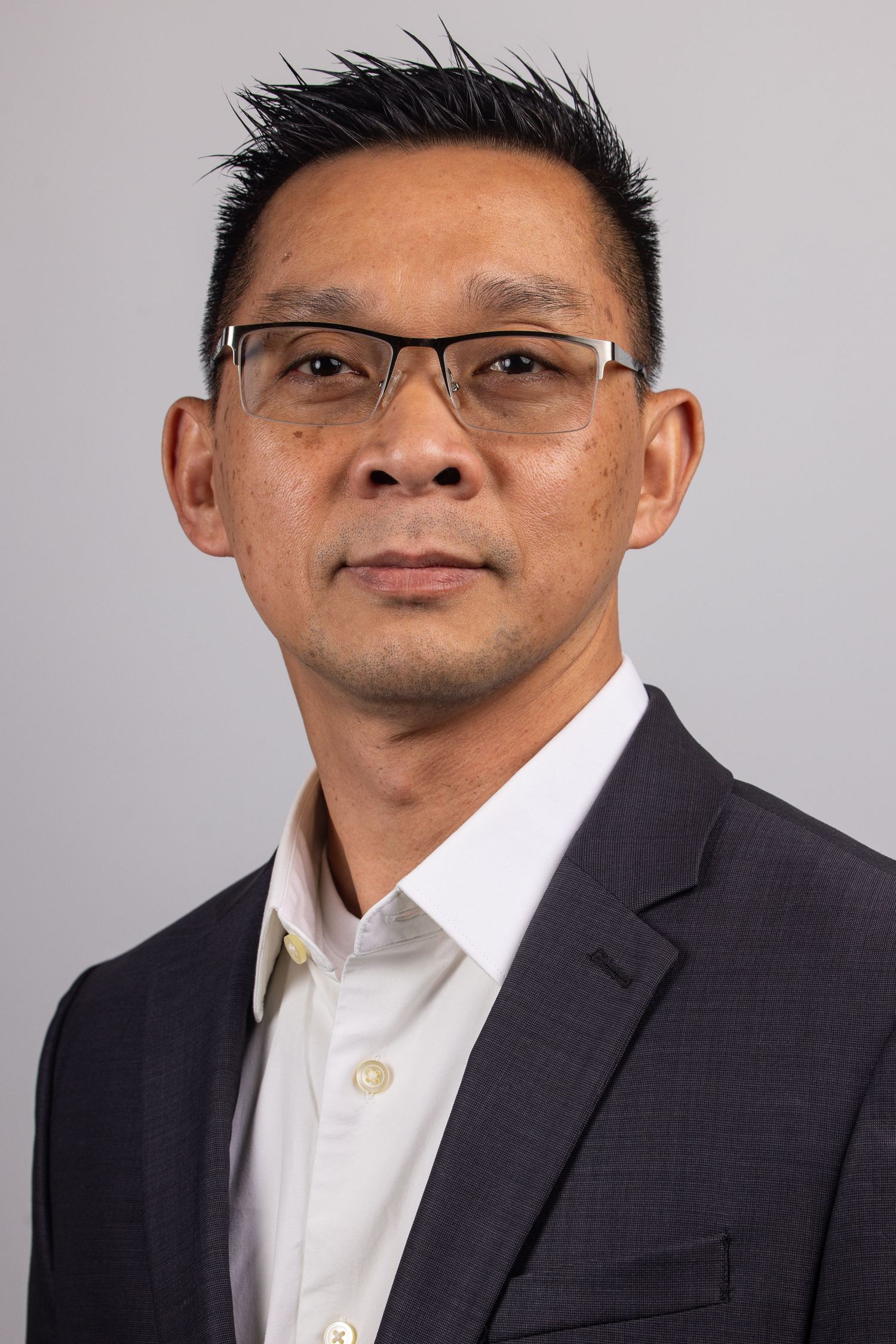 Professional headshot of an Asian man wearing glasses and a dark blazer with white shirt against a gray background.