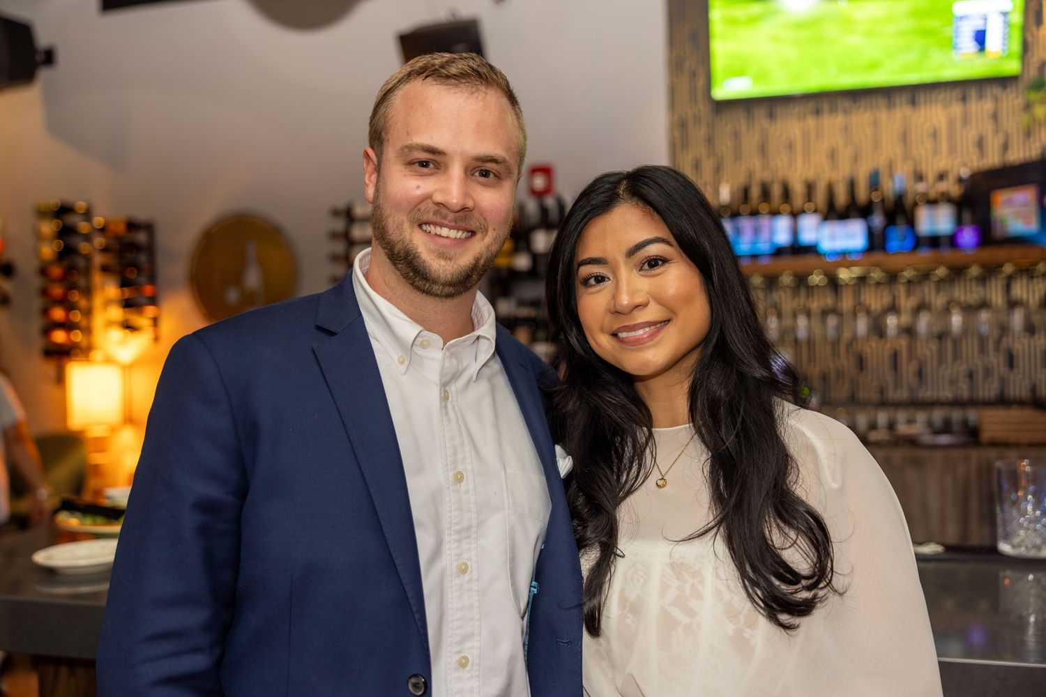 A smiling couple poses together in a stylish bar restaurant setting, man in blue blazer and woman in white top.