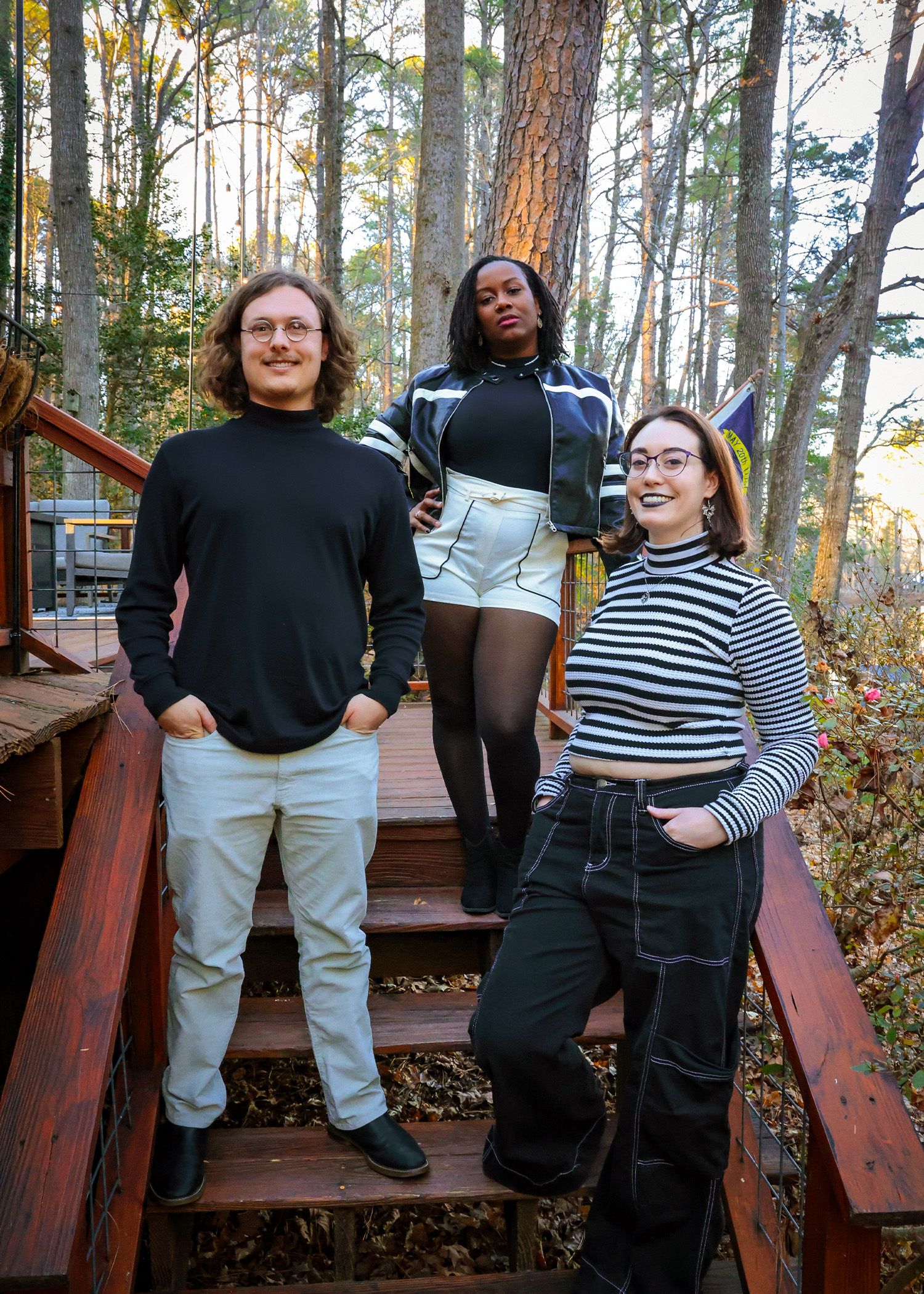 Three friends in casual outfits pose together on wooden stairs in a forest setting.