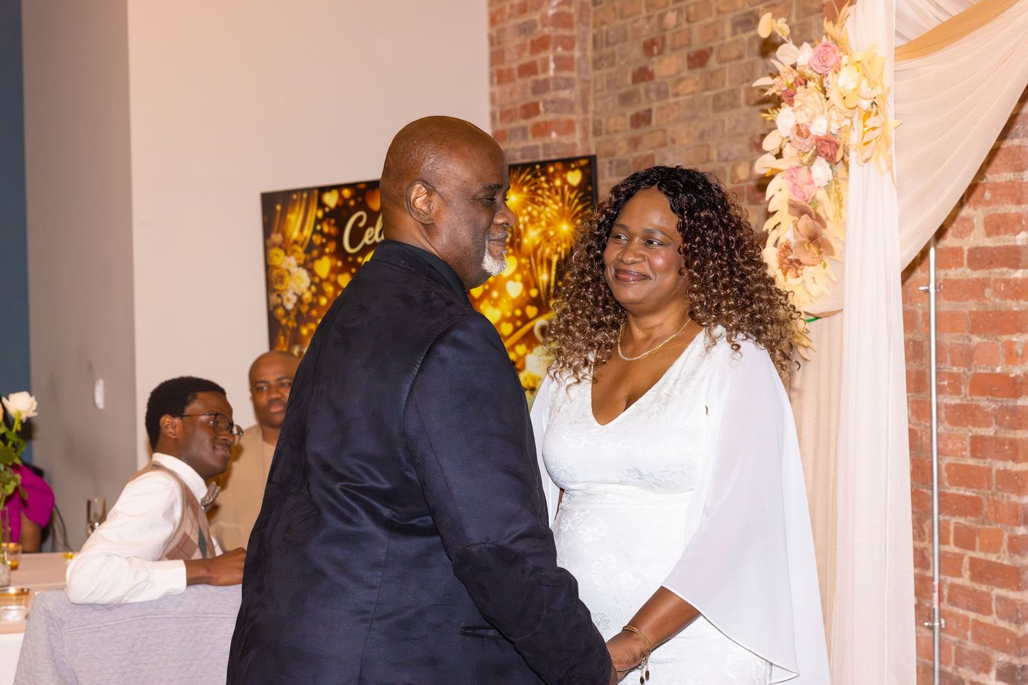 A couple shares a tender moment during their wedding ceremony in a decorated brick-walled venue.
