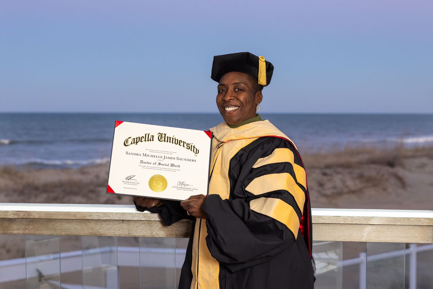Graduate in cap and gown proudly holds Capella University diploma outdoors near ocean backdrop.