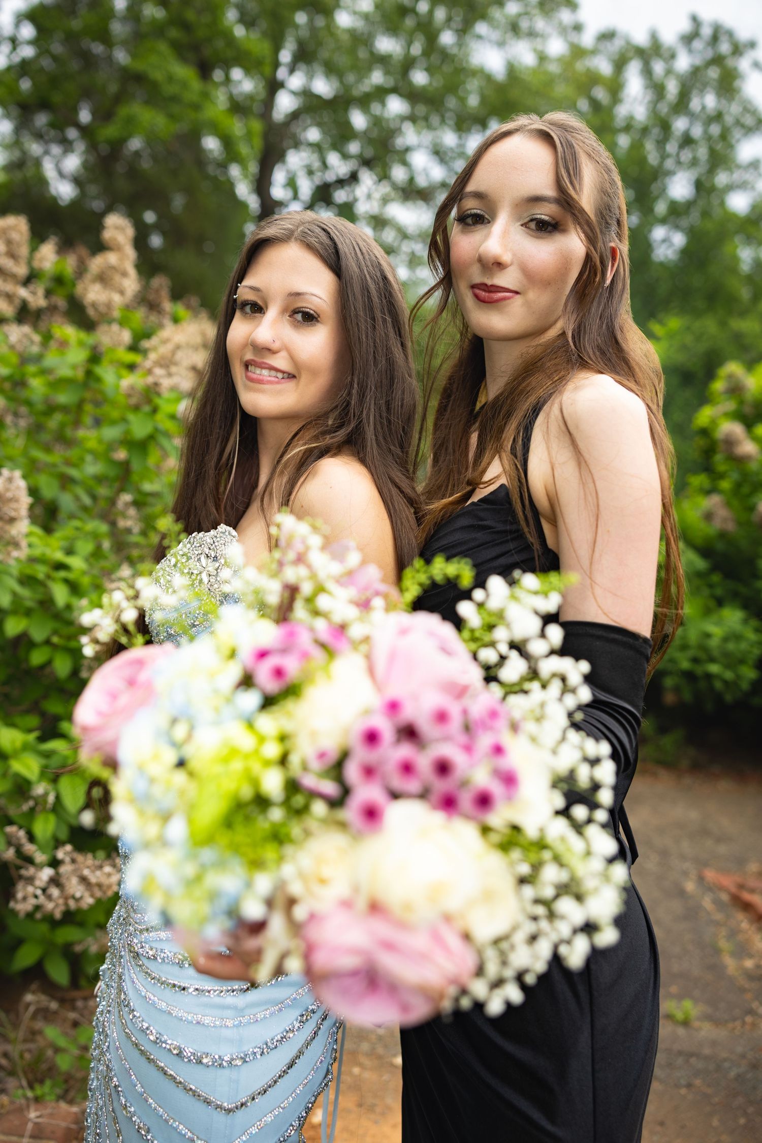Two young women in elegant prom dresses pose outdoors holding a bouquet of pink and white flowers.