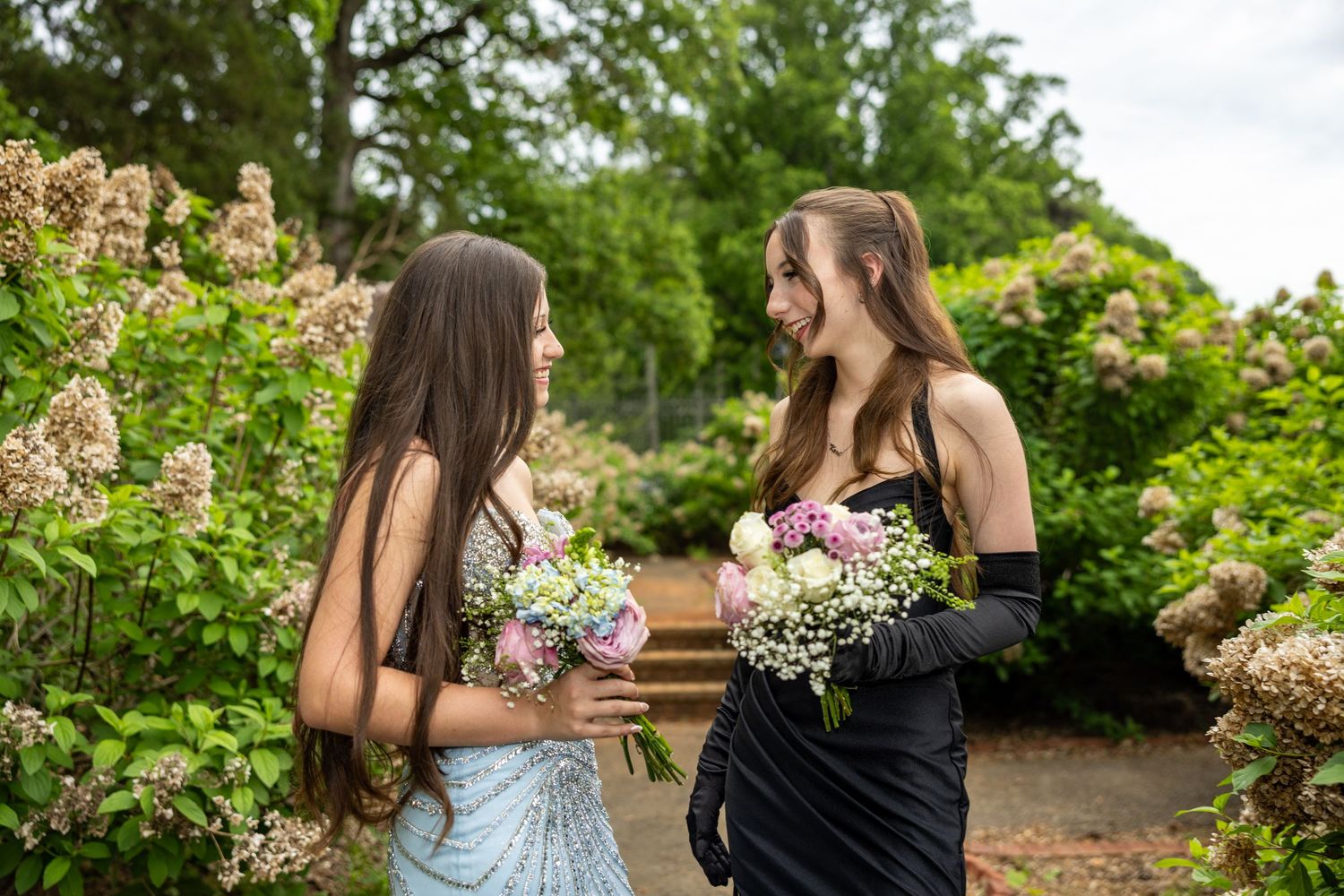 Two teenage girls in formal gowns holding bouquets smile at each other in a lush garden with blooming hydrangeas.