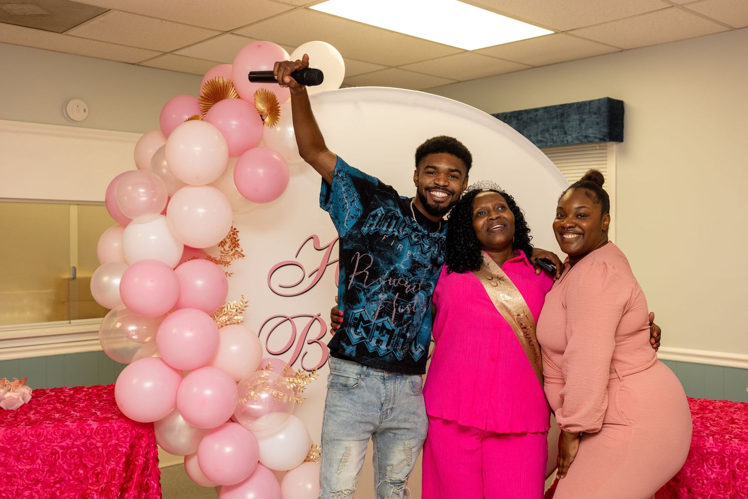 Three people posing and smiling at a pink-themed baby shower with balloon arch decorations.