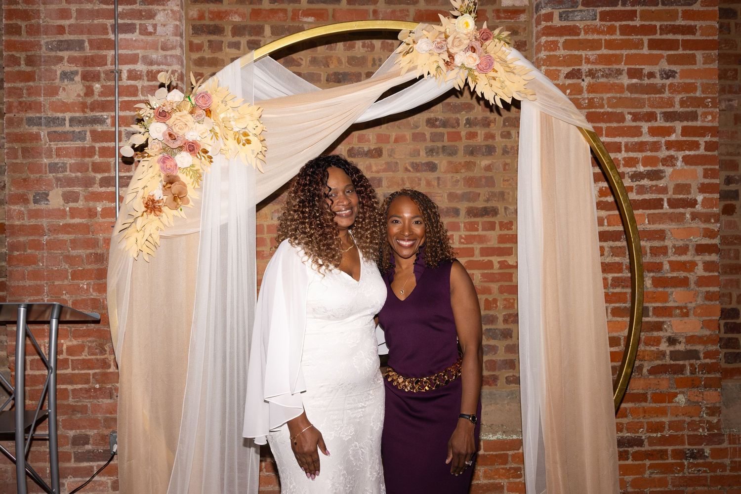 Two women smiling under a gold circular arch with floral decor and white draping at an elegant indoor venue.