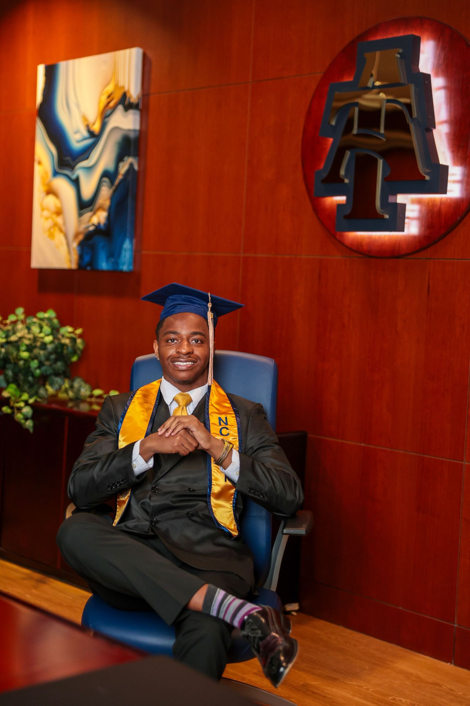 Graduate in black cap and gown with gold stole sits in blue chair against red wall.