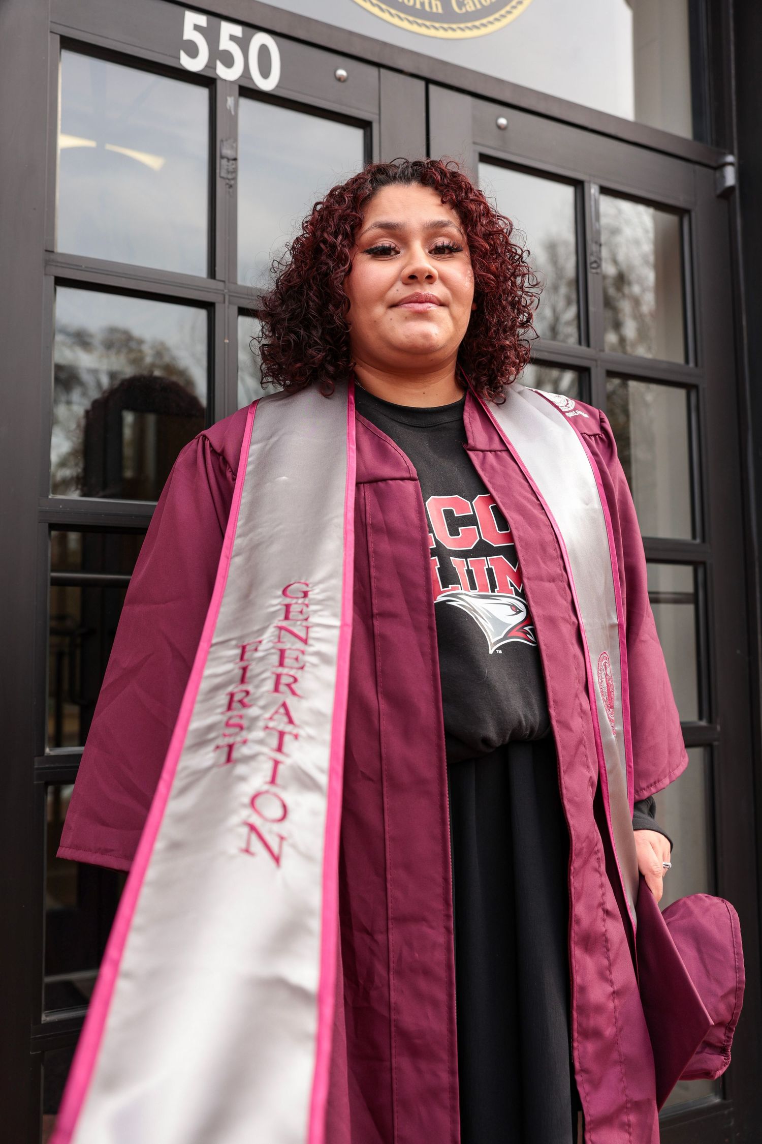 Graduate in maroon academic regalia with white sash stands in front of building entrance.