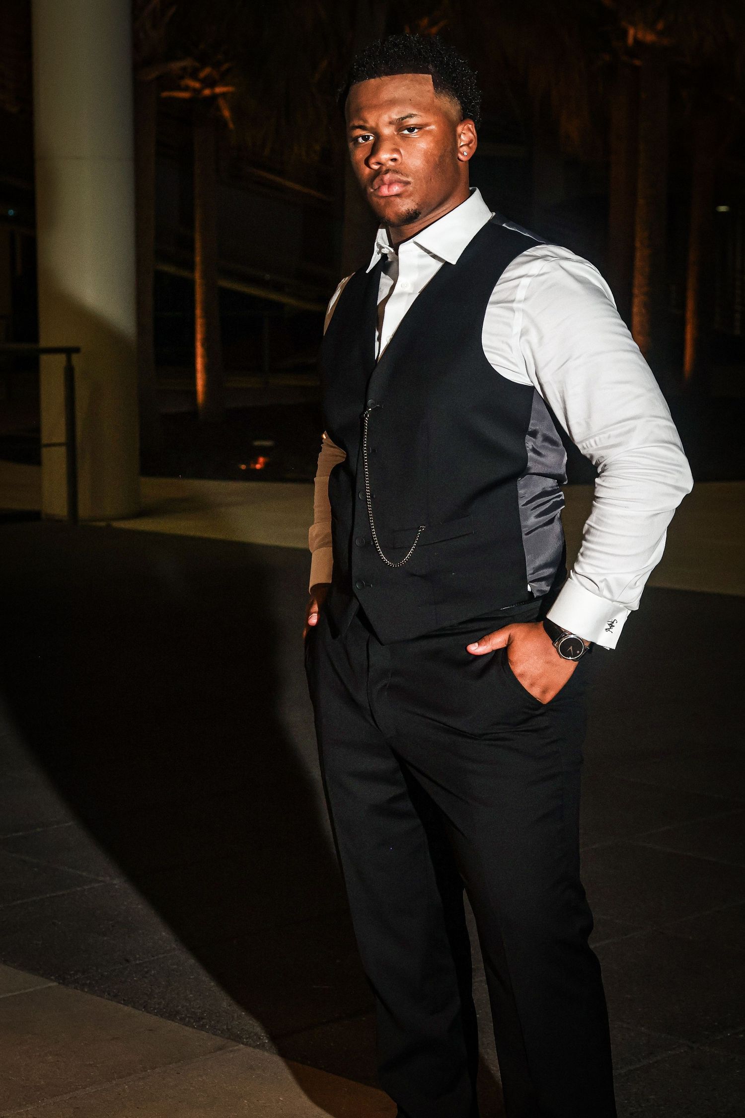 A stylish formal portrait features someone in a black vest and white shirt against a dark background.