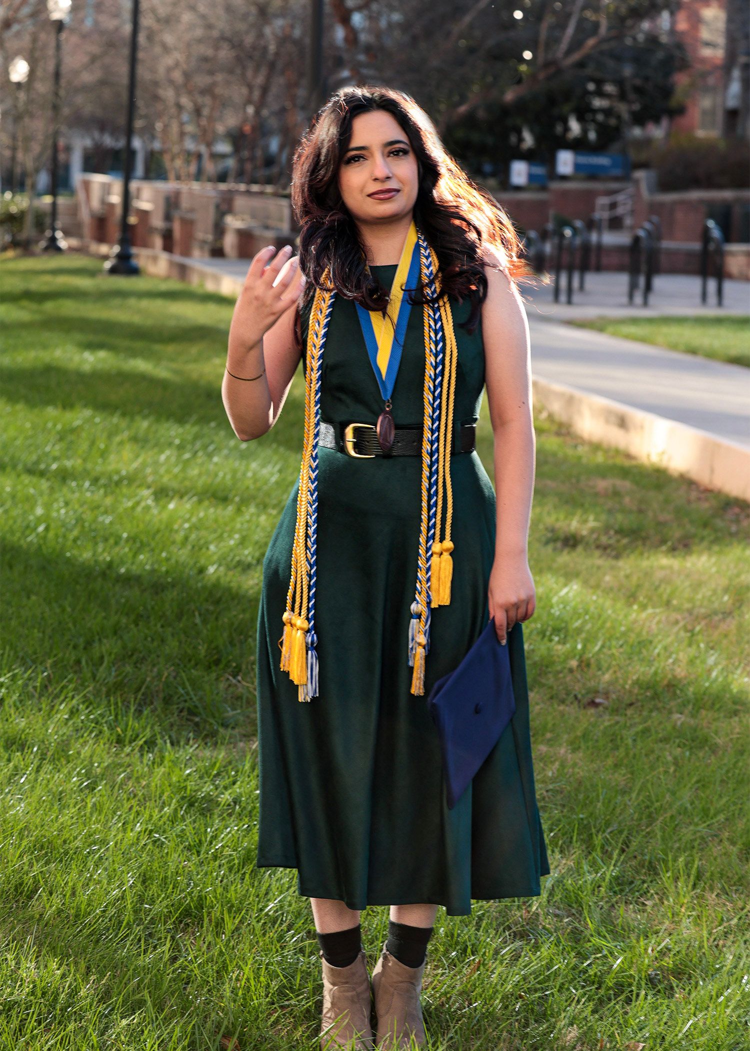 Graduate in green academic dress with gold cords poses on grassy campus hillside.