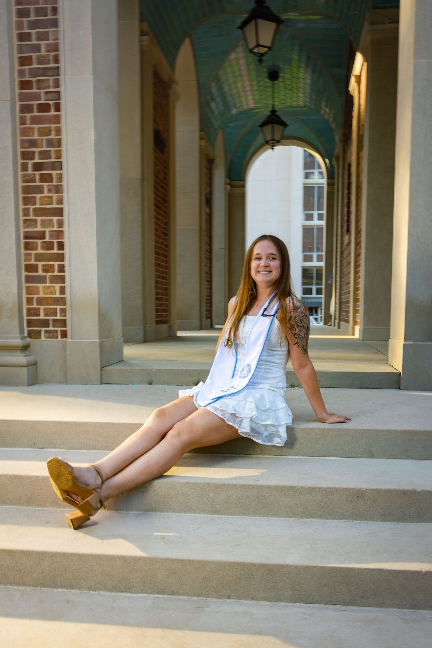 Young woman in white dress and yellow heels sitting on stone steps of a brick building with arched colonnade.