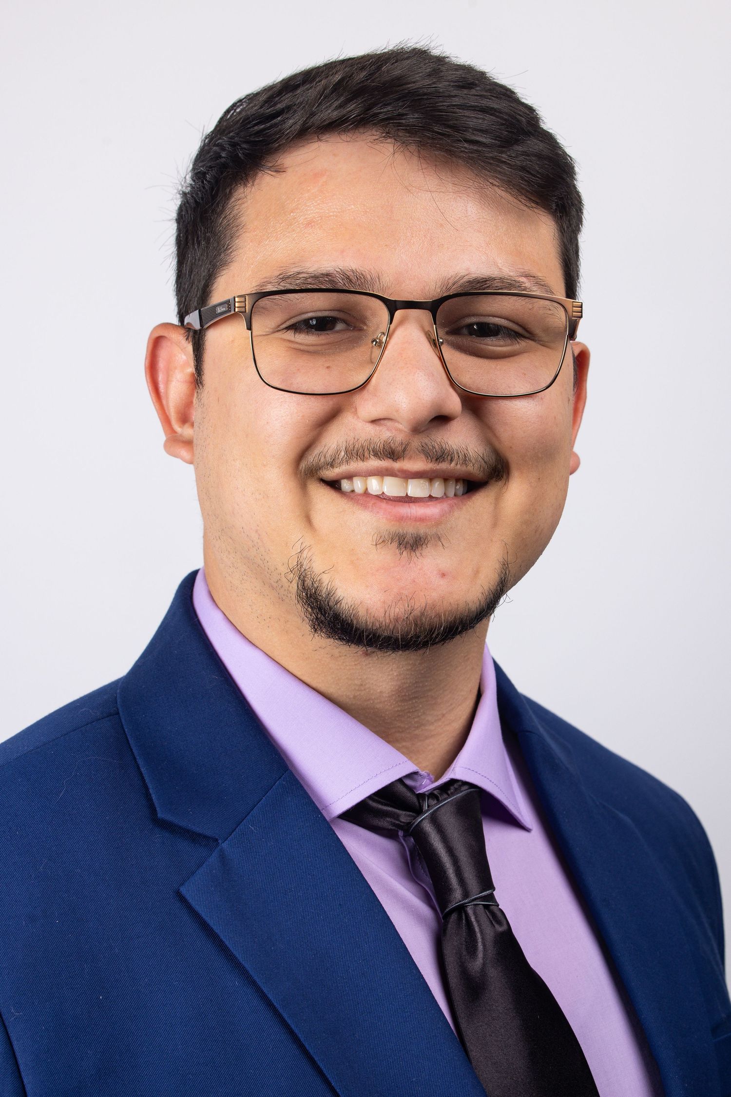 Professional man smiling in blue suit, purple shirt, black tie, and glasses against light background.
