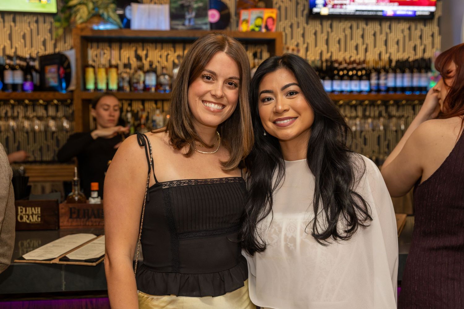 Two women smiling together at a bar, one in a black crop top and one in a white blouse, with shelves of bottles behind them.