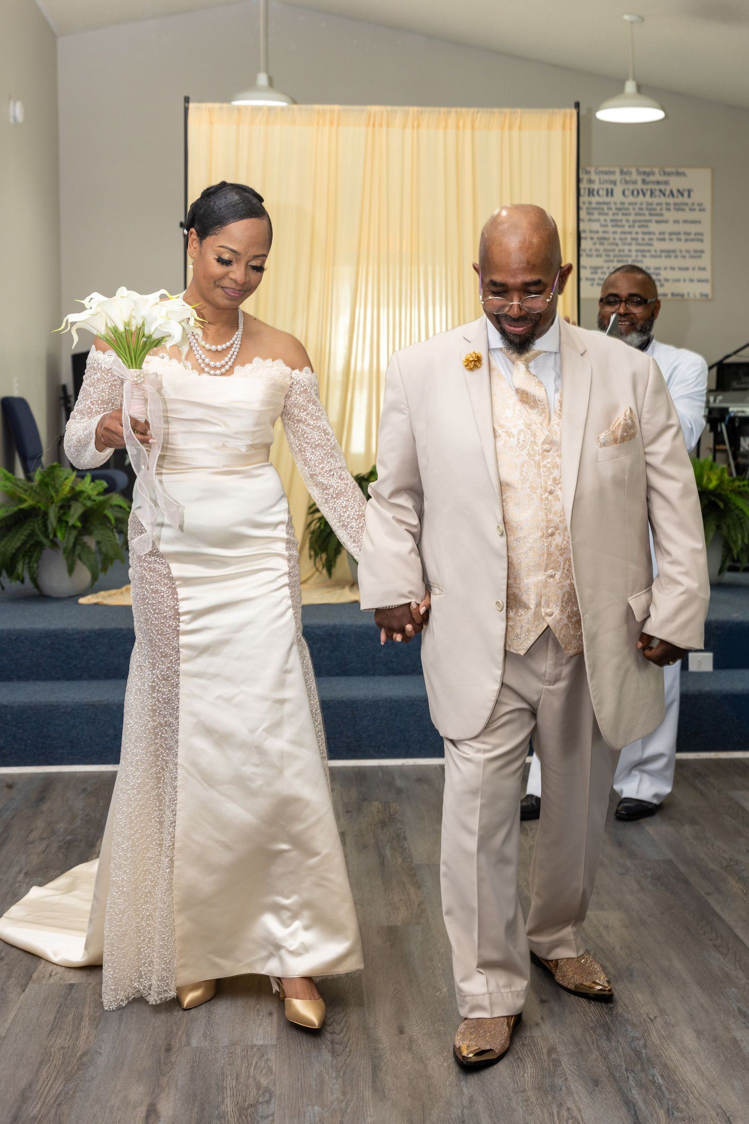 Bride and groom in elegant ivory wedding attire walk hand in hand down the aisle in a church ceremony.