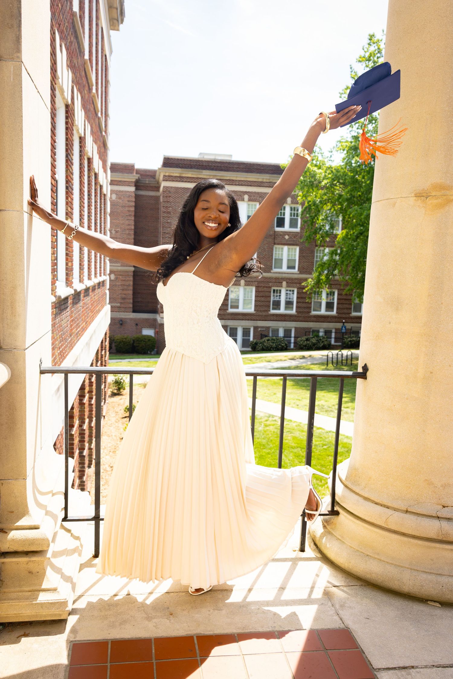 Young Black woman in white dress celebrates graduation, holding cap with arms raised, smiling on university campus steps.