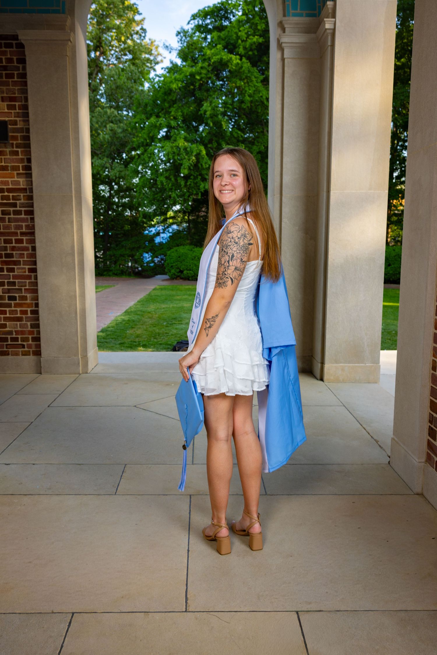 Young woman in white dress and blue graduation gown holding cap, smiling between brick columns outdoors.