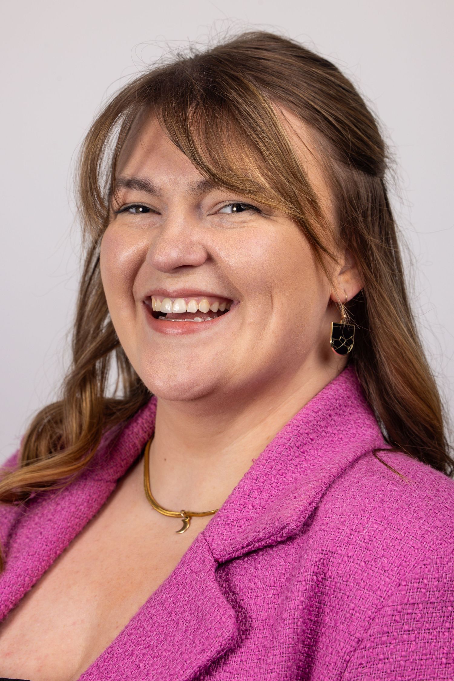 Smiling woman with brown hair wearing a pink blazer and gold necklace poses for a professional headshot.