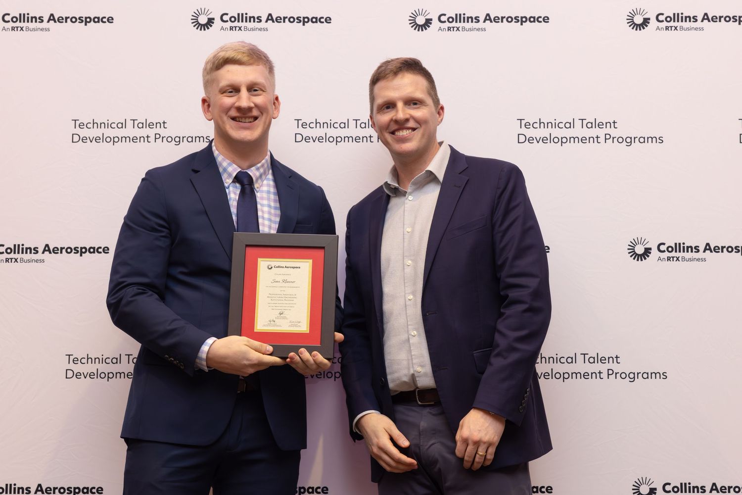 Two men pose together at a Collins Aerospace event, one holding a framed award certificate.