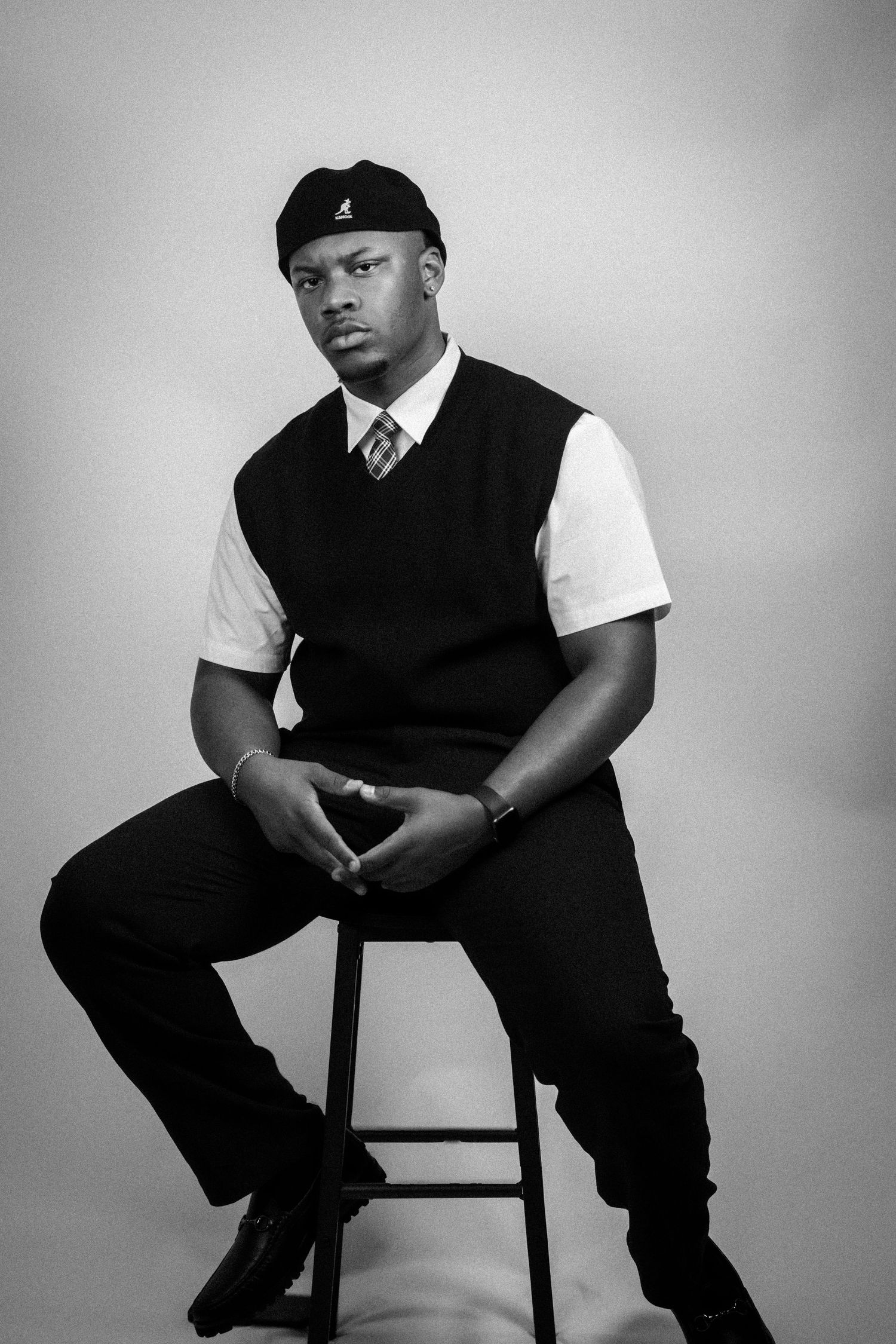 Black and white portrait of a young man in a vest, tie, and cap sitting on a stool in a studio.